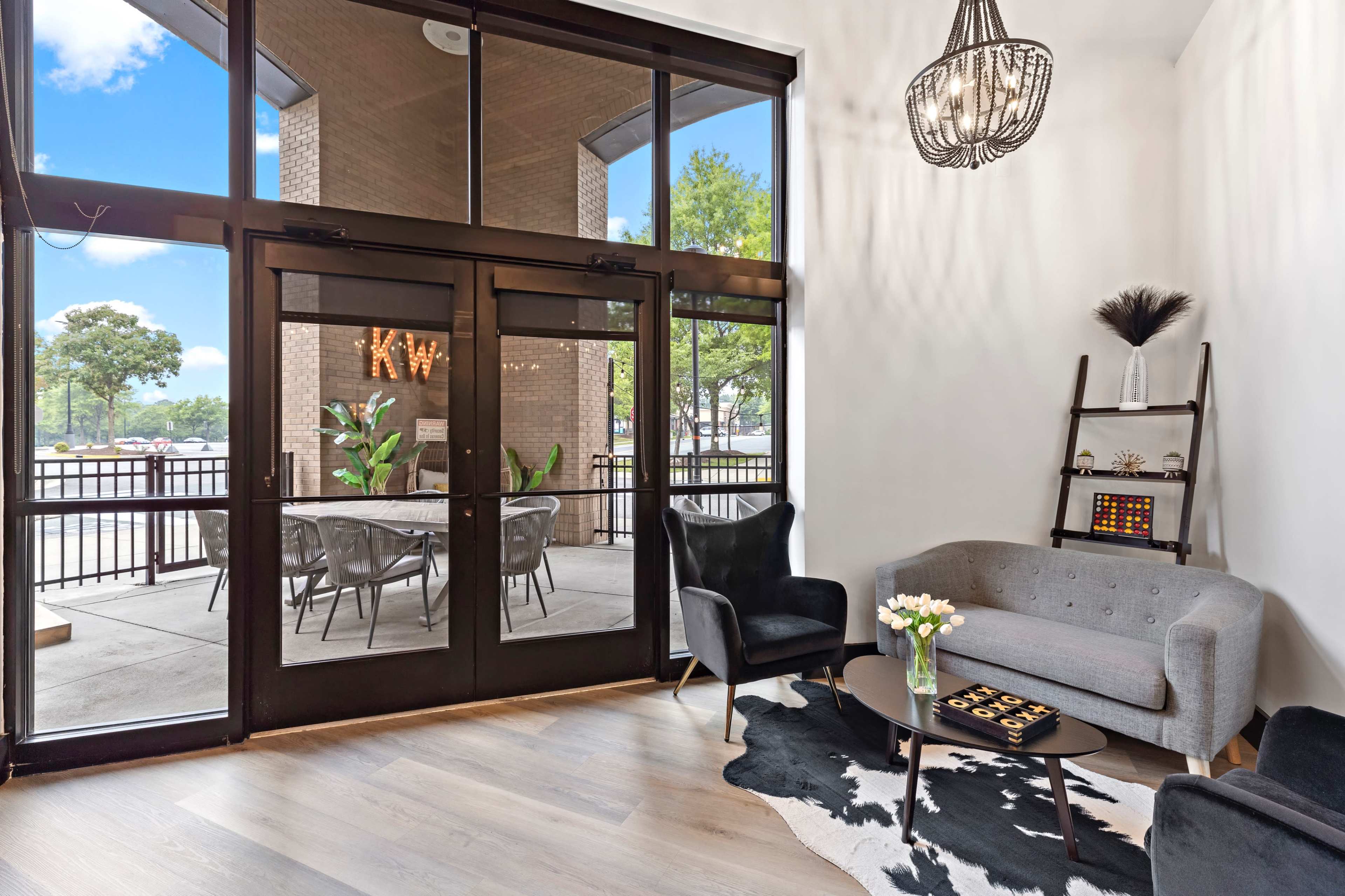 A modern lobby features large glass doors leading to an outdoor seating area, a gray sofa, black chairs, and decorative elements on a wooden shelf.
