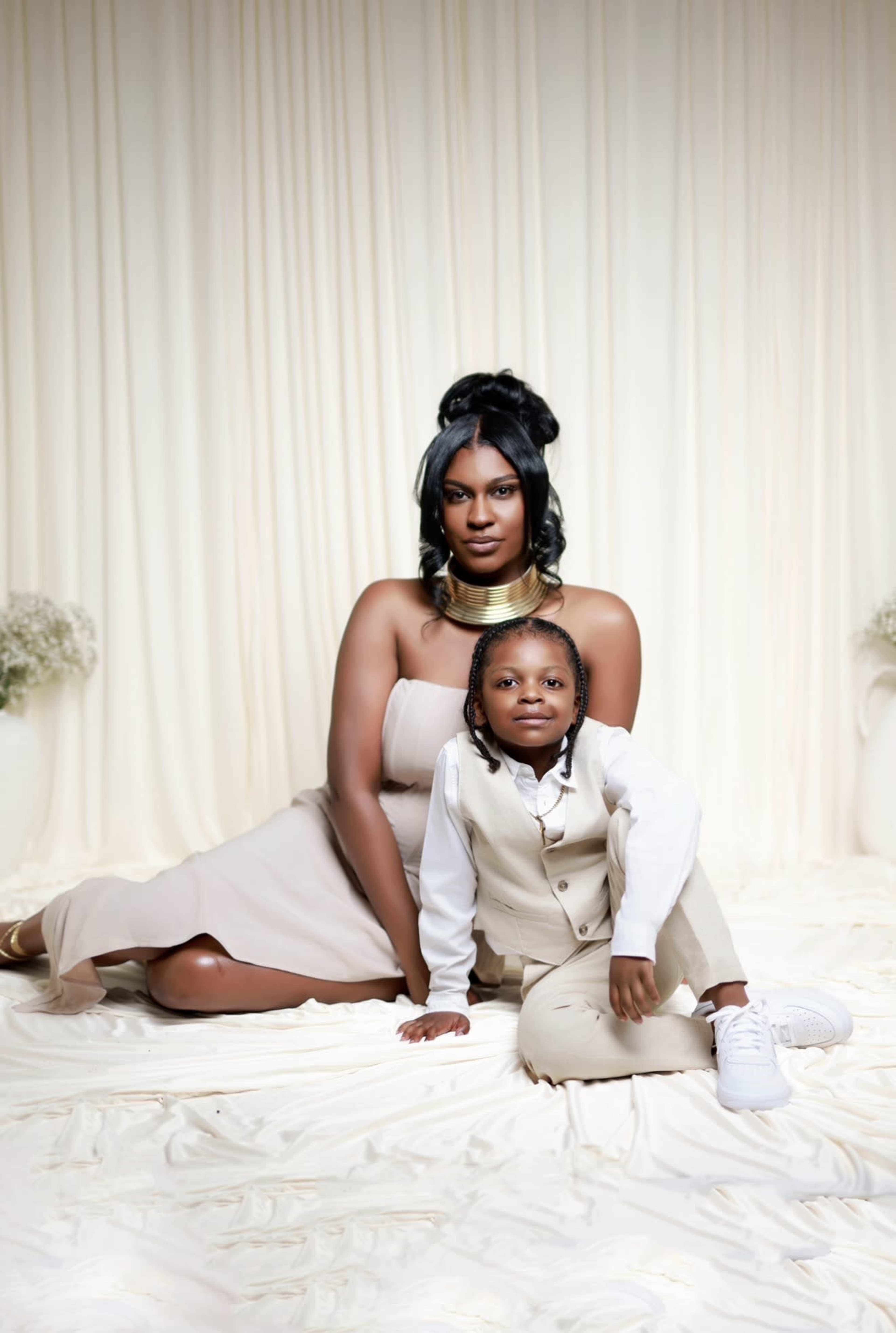 A woman sits gracefully on a draped surface while a child kneels beside her, both wearing stylish outfits against a soft, neutral backdrop.