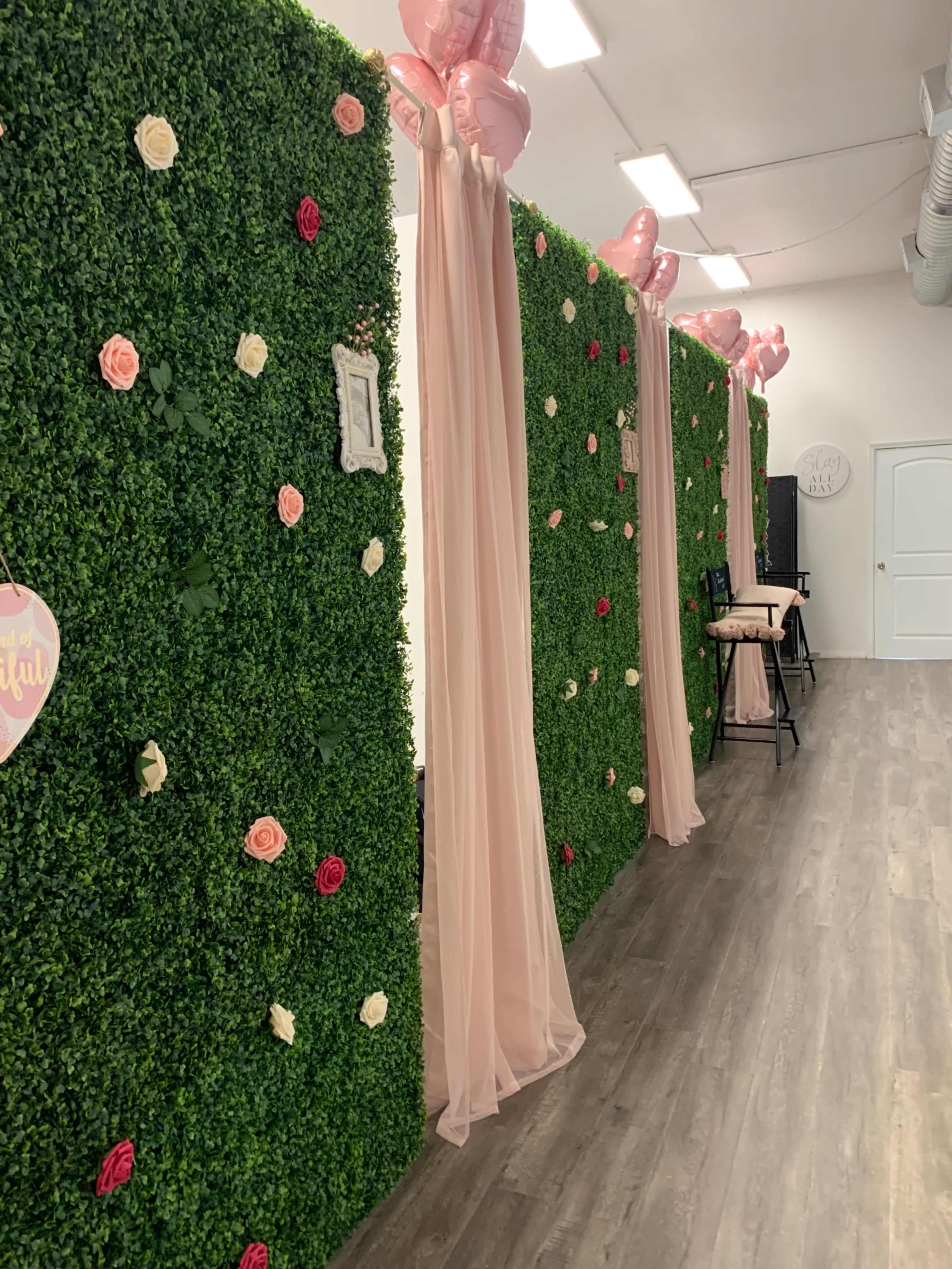 A decorated interior space with artificial green foliage walls, draped fabric, and pink balloons.