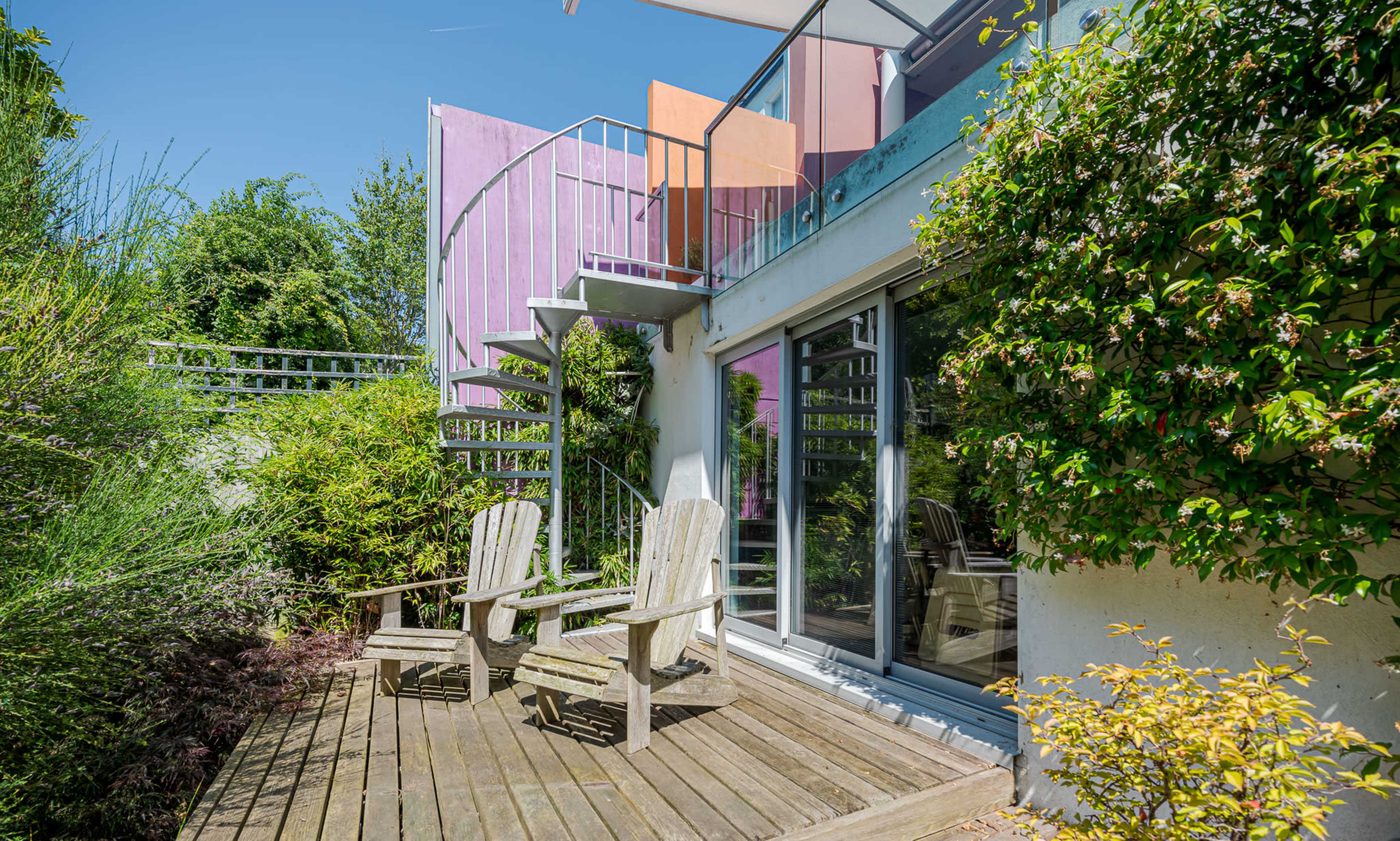 The image shows a modern house with a vibrant purple accent wall, surrounded by greenery and featuring a spiral staircase leading to an upper balcony, with two wooden chairs situated on a deck.