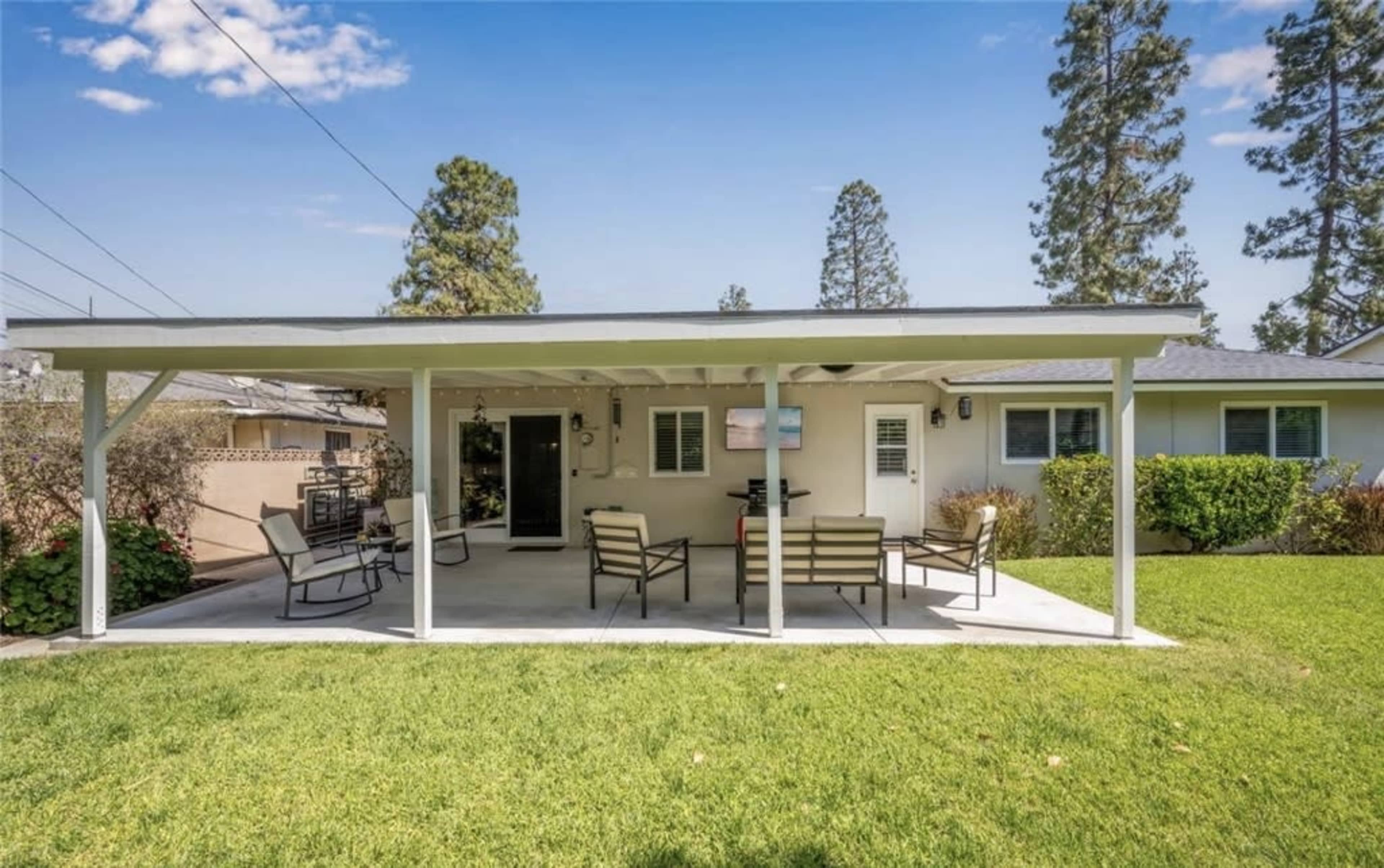 The image shows a patio area in front of a house, featuring a covered seating arrangement with chairs and a table on a green lawn.