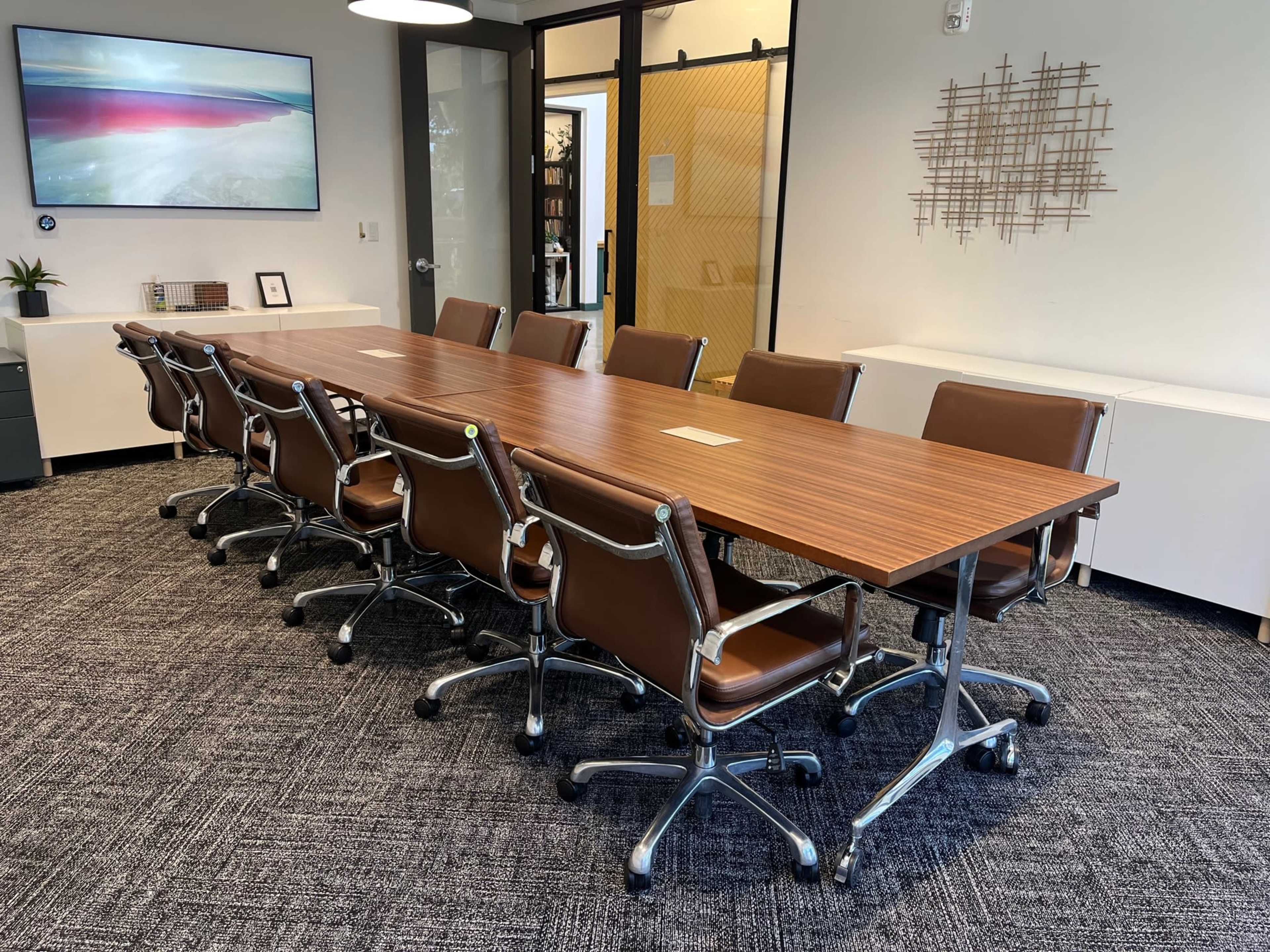 A long wooden conference table with brown leather chairs is set up in a modern office meeting room.