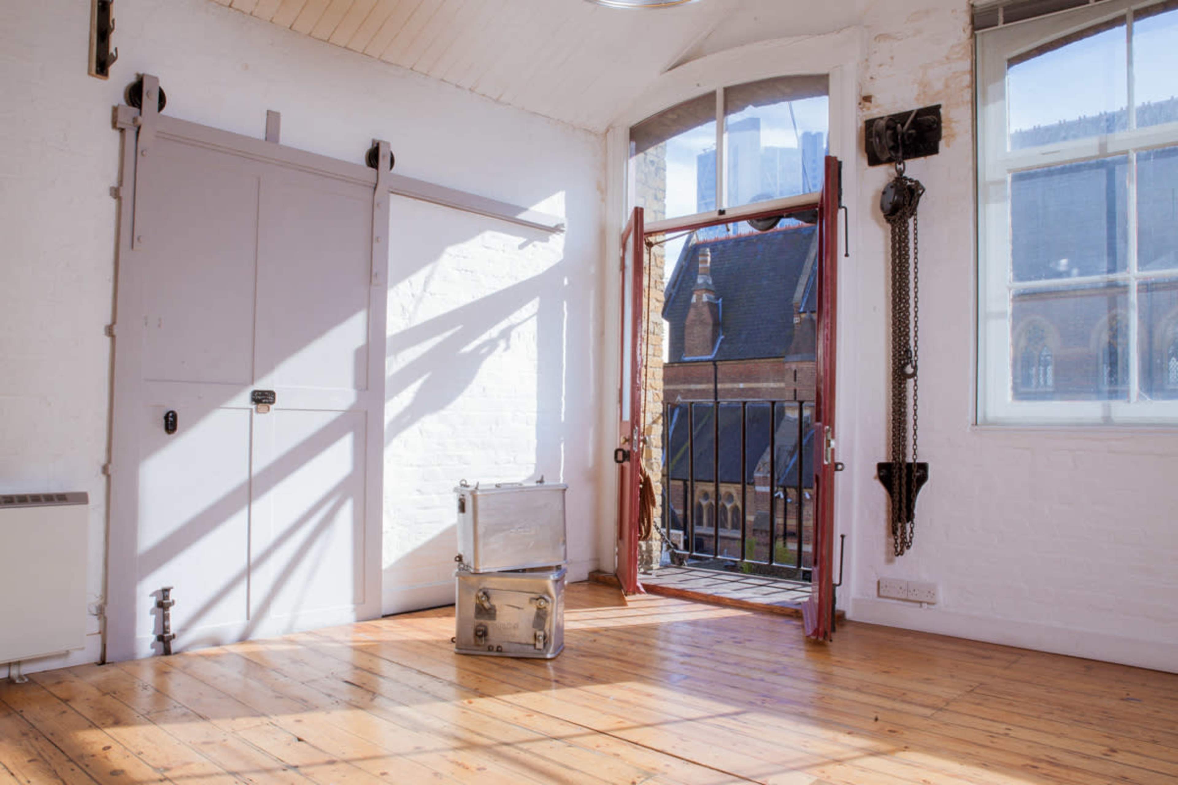 A bright, empty room with wooden floors, large windows, and an open door leading to a balcony, featuring two metallic suitcases on the floor.