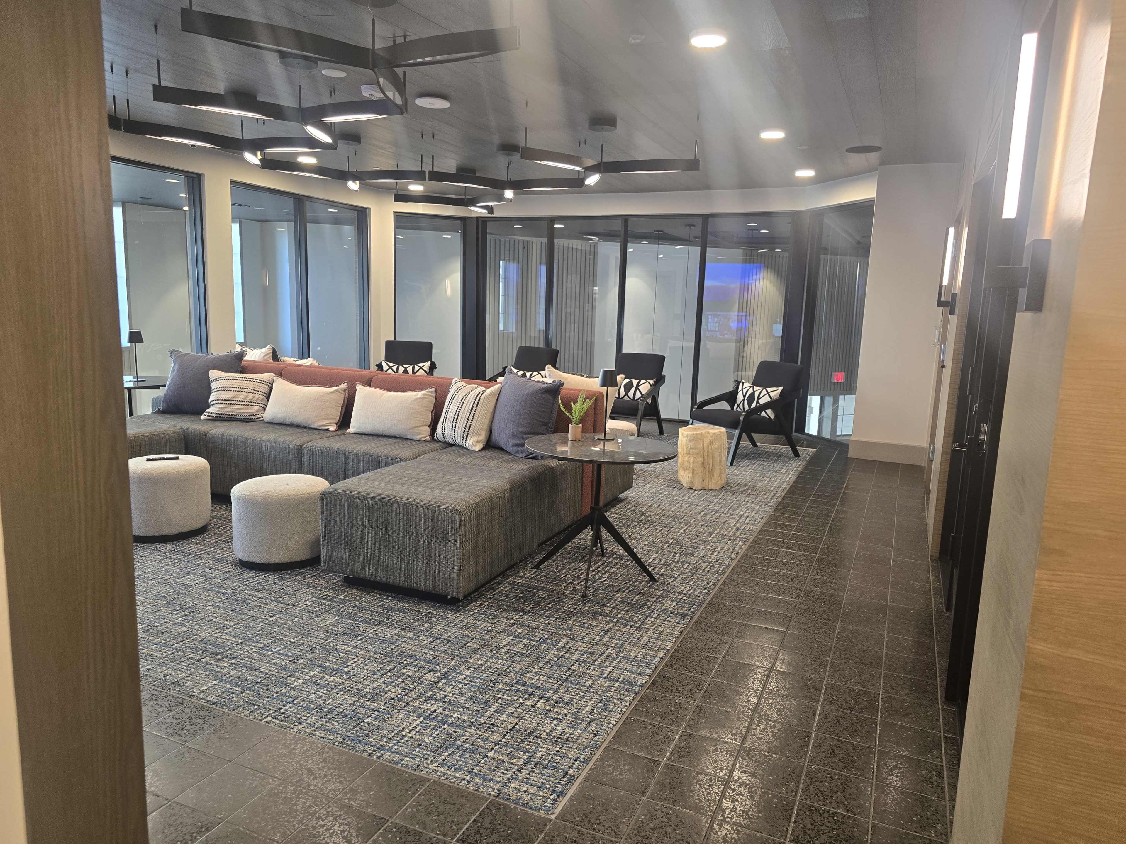 The image shows a modern lounge area featuring a large sectional sofa, round ottomans, a coffee table, and several ceiling fans, all within a glass-walled space.