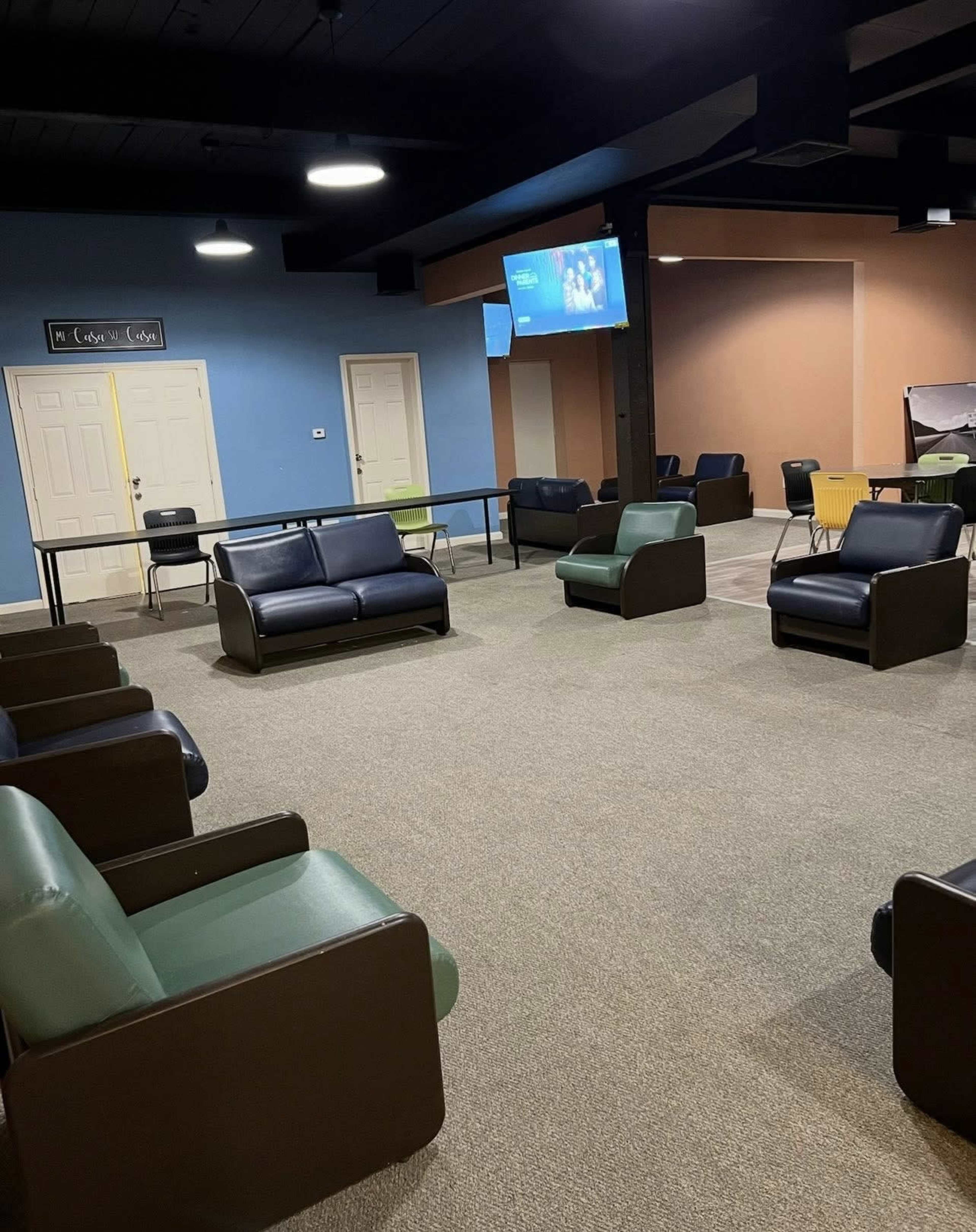 The image shows a lounge area with multiple chairs arranged around a central open space, and two screens mounted on the walls.
