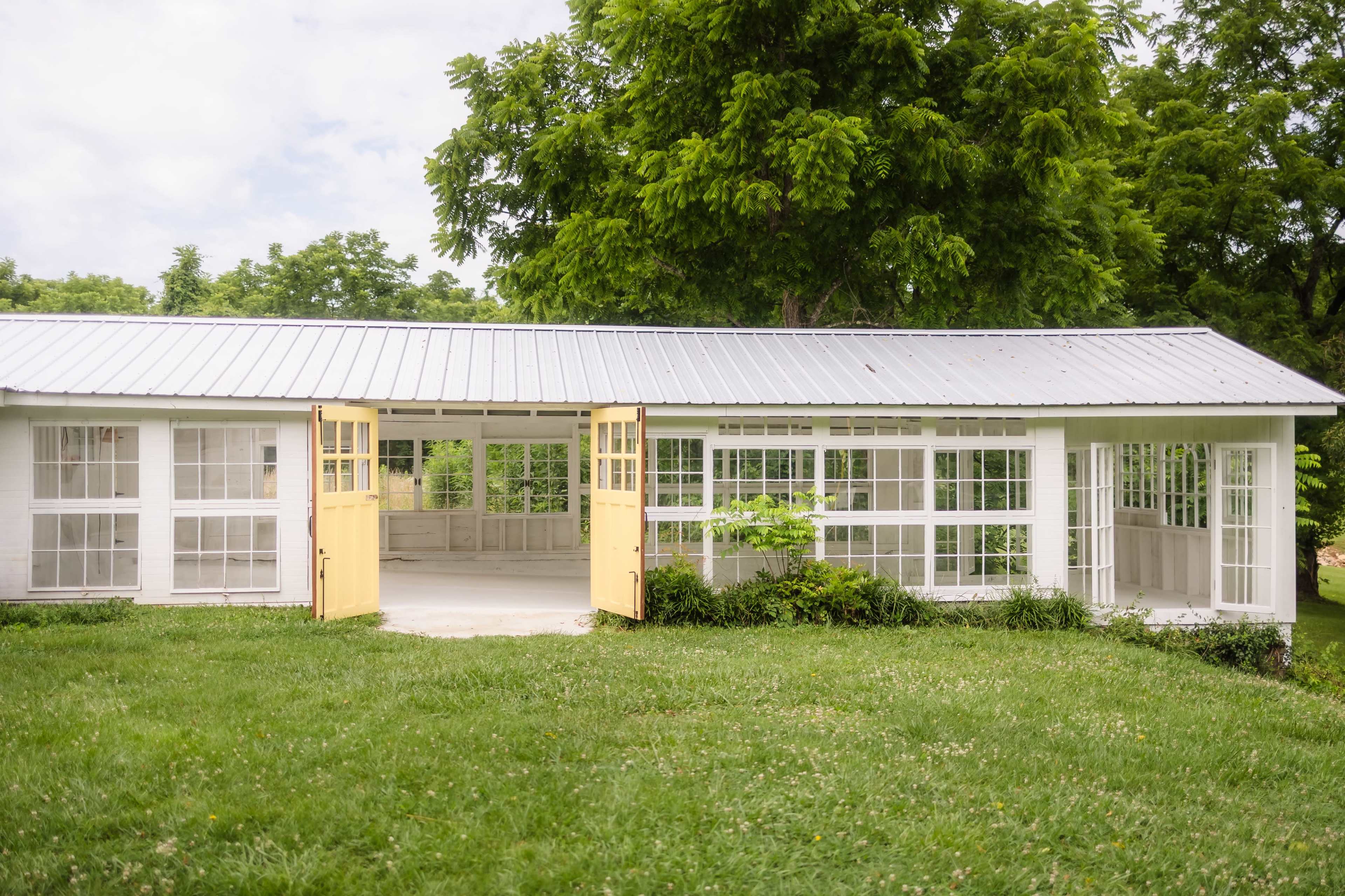 The image shows a white, open-air structure with large glass windows and yellow double doors, surrounded by green grass and trees.
