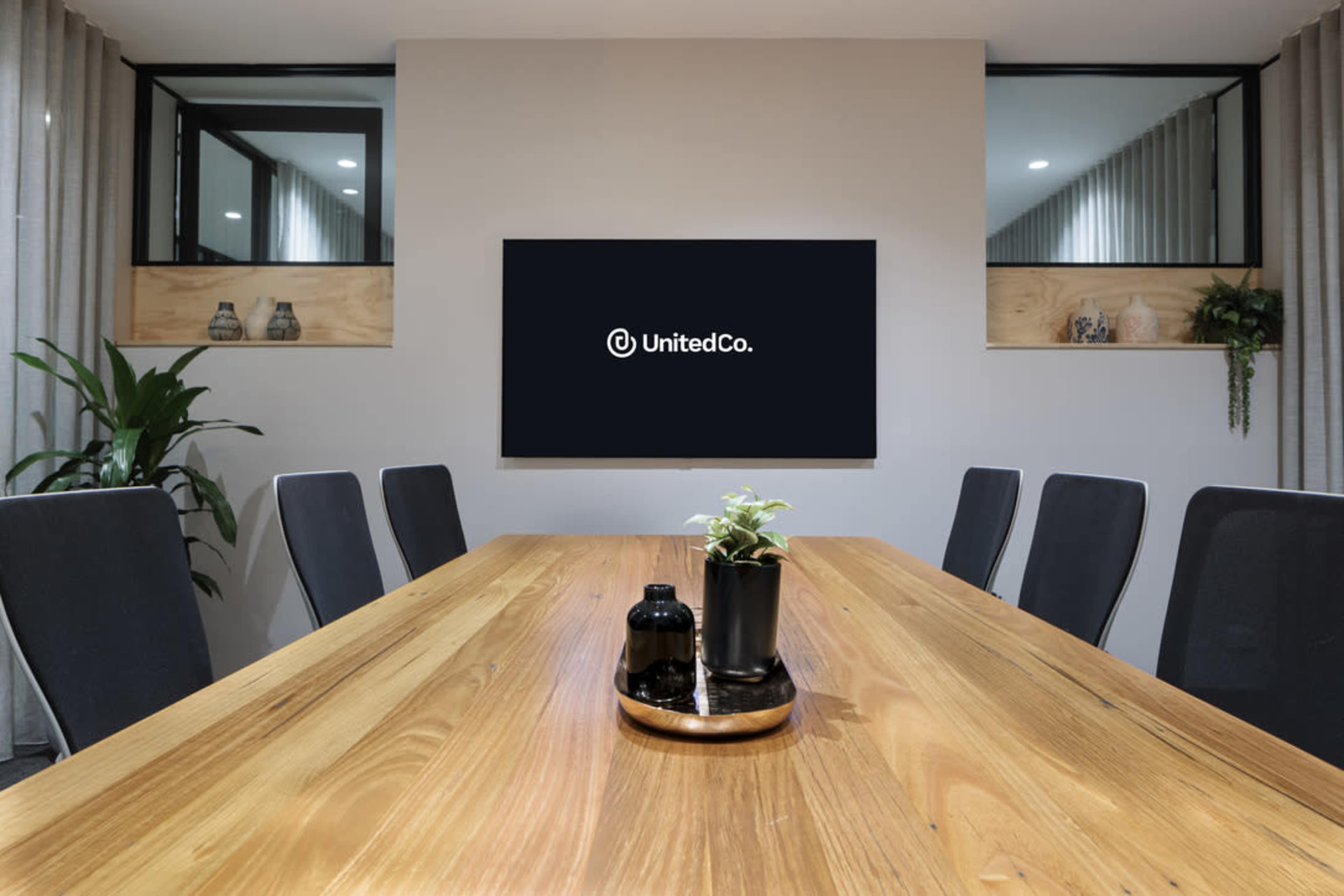 A modern conference room features a large wooden table surrounded by black chairs, with a television displaying the UnitedCo logo on the wall.