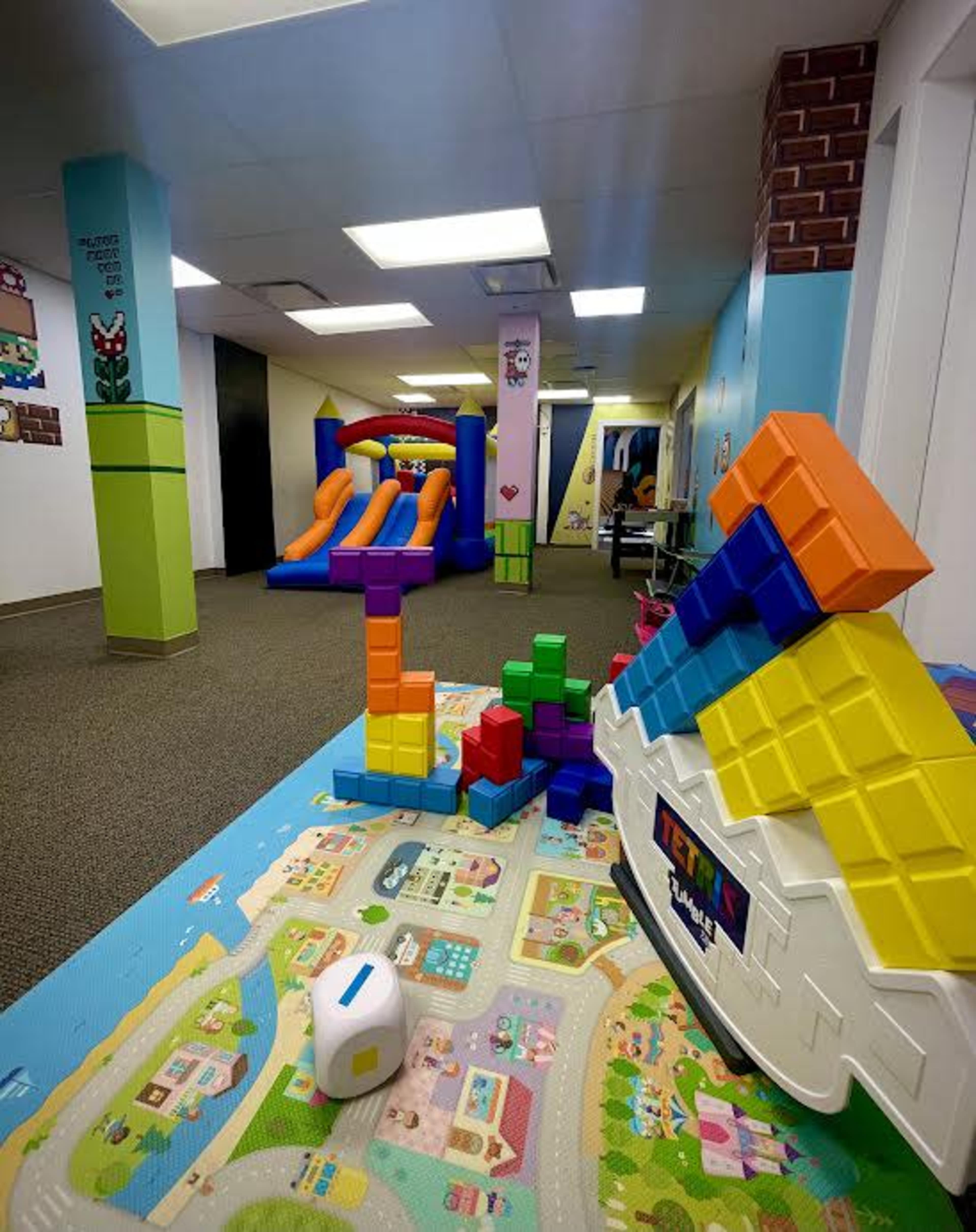 The image shows an indoor play area featuring colorful soft play structures, oversized building blocks, and a colorful play mat with cityscape designs.