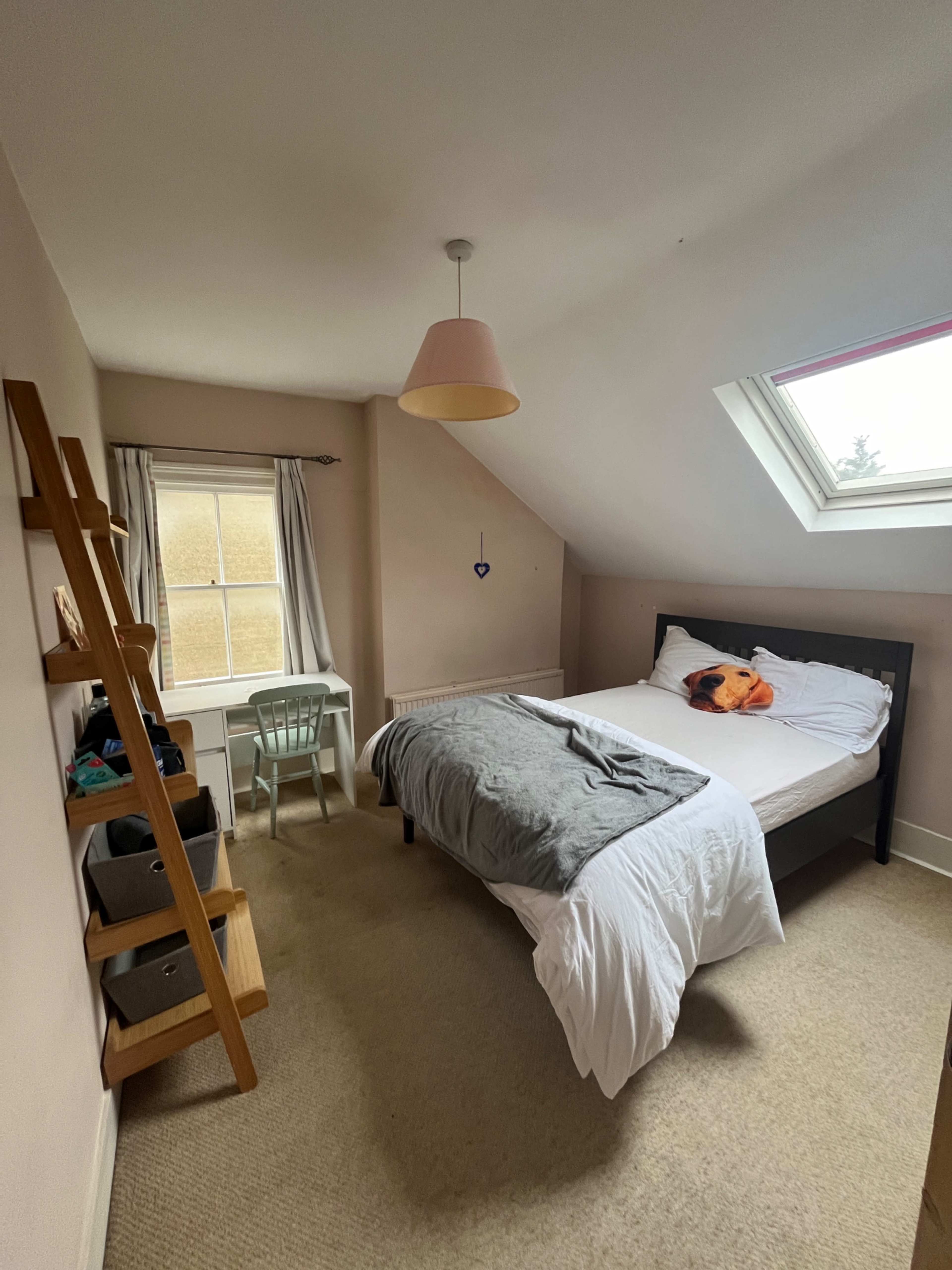 The image shows a simple bedroom with a bed, a small desk and chair, a ladder shelf, and a window with a skylight.