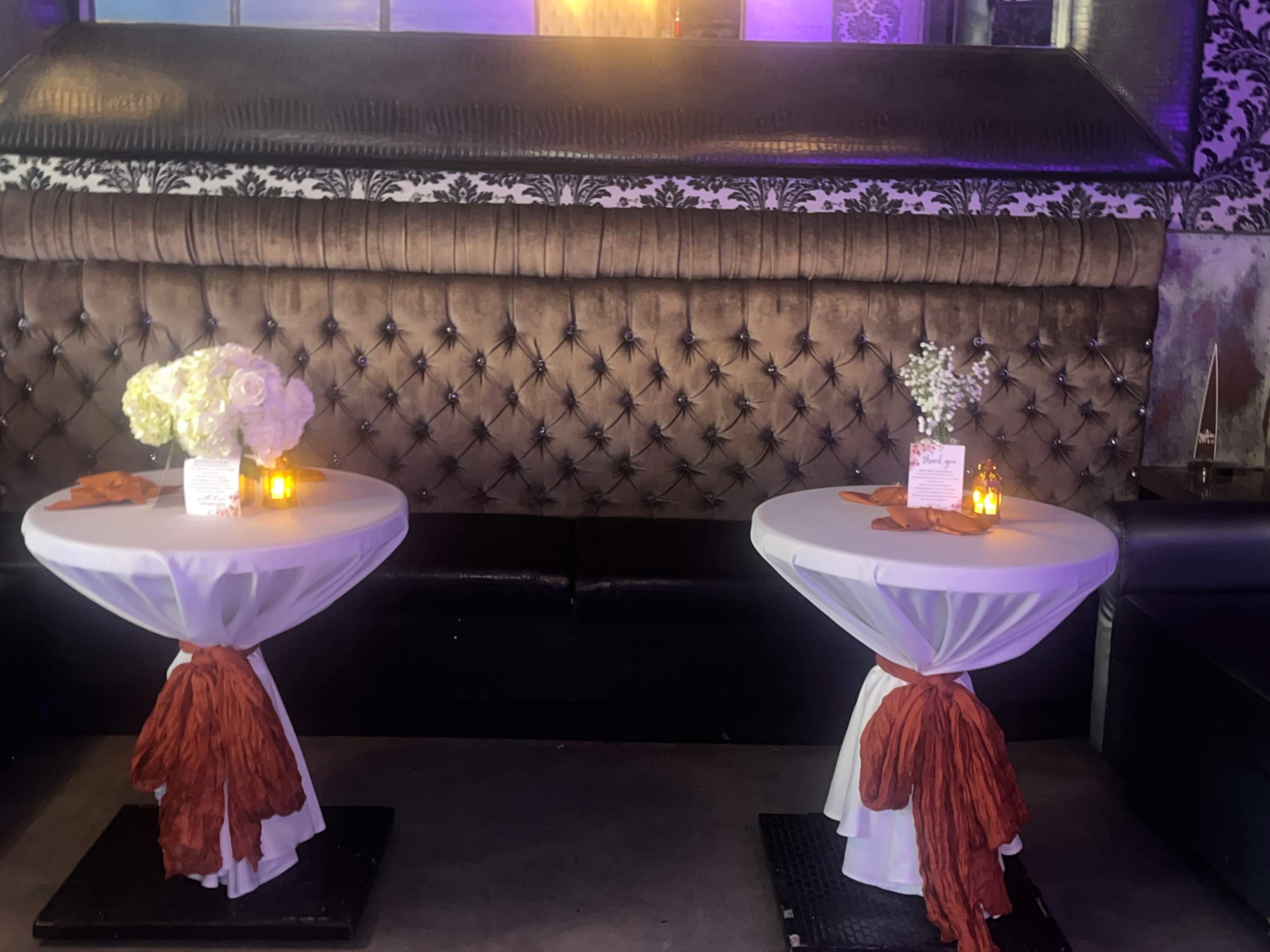 Two round tables with white tablecloths and orange sashes are set up in front of a tufted brown leather backdrop, with floral arrangements and candles on each table.