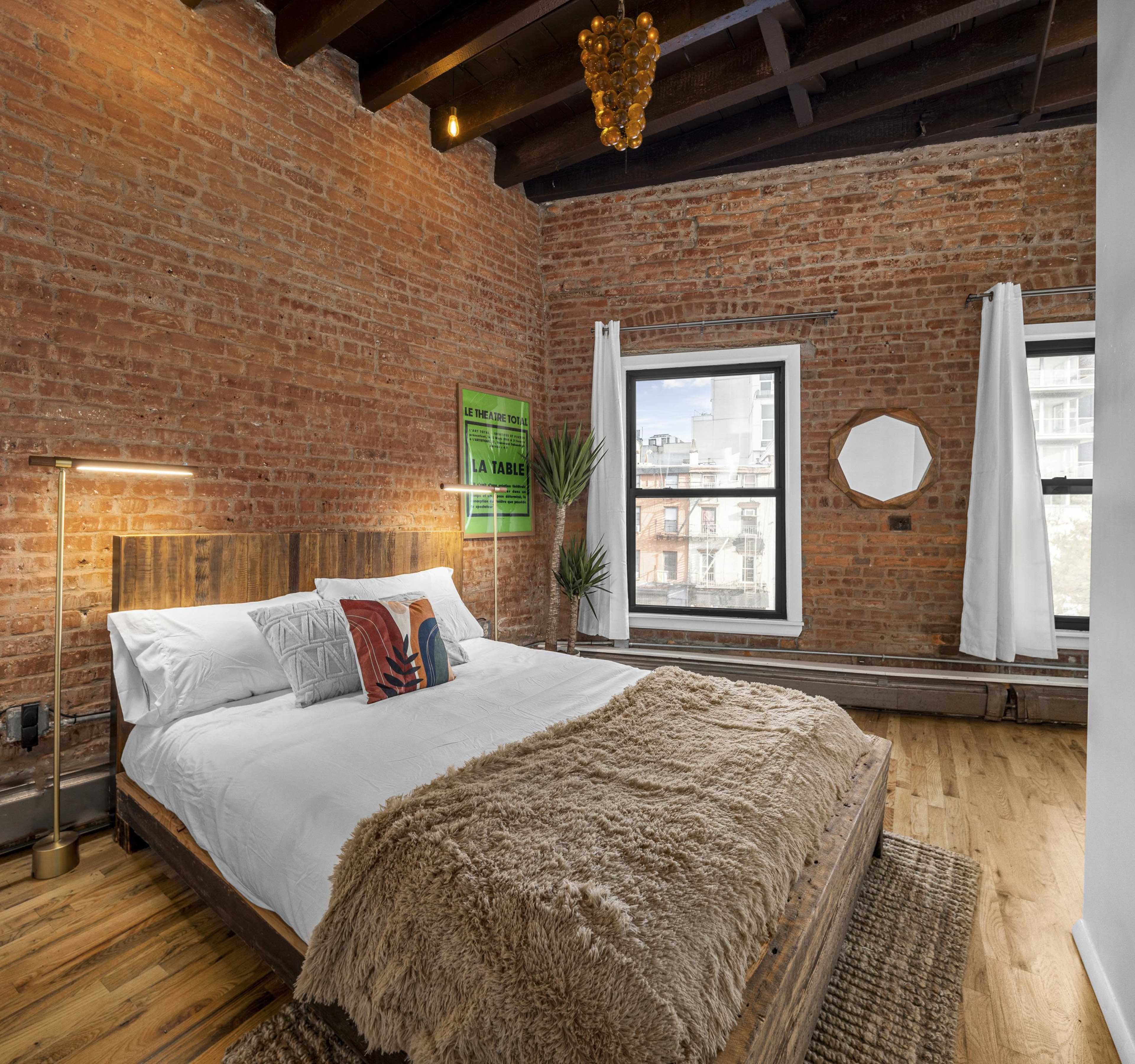 A modern bedroom with a brick wall, a wooden bed frame covered in white bedding, and large windows allowing natural light to enter.