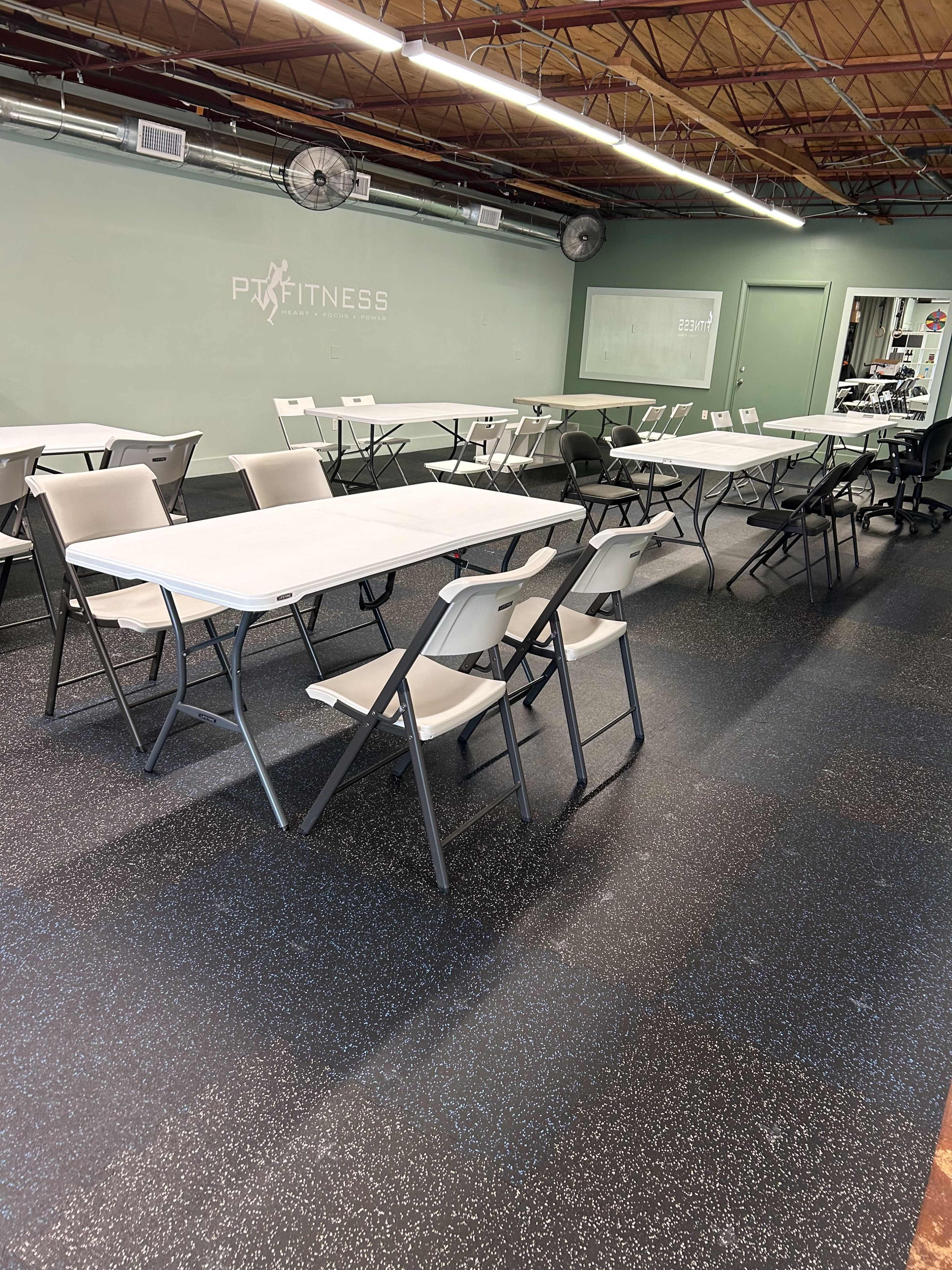 A fitness studio with several rows of white folding tables and chairs set up on a black rubber floor.