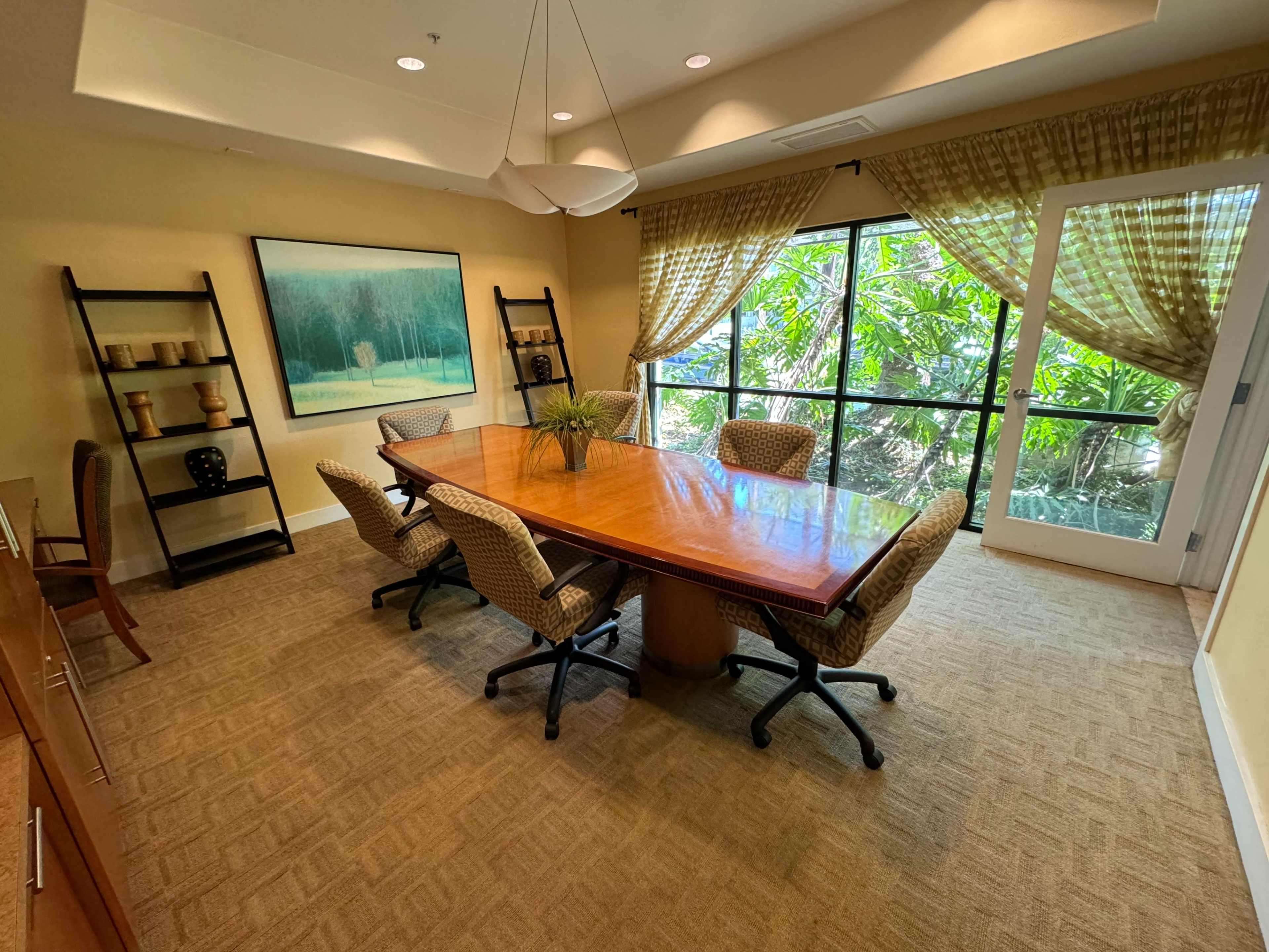 A conference room features a large wooden table surrounded by six chairs, with a window view of greenery and artwork on the wall.
