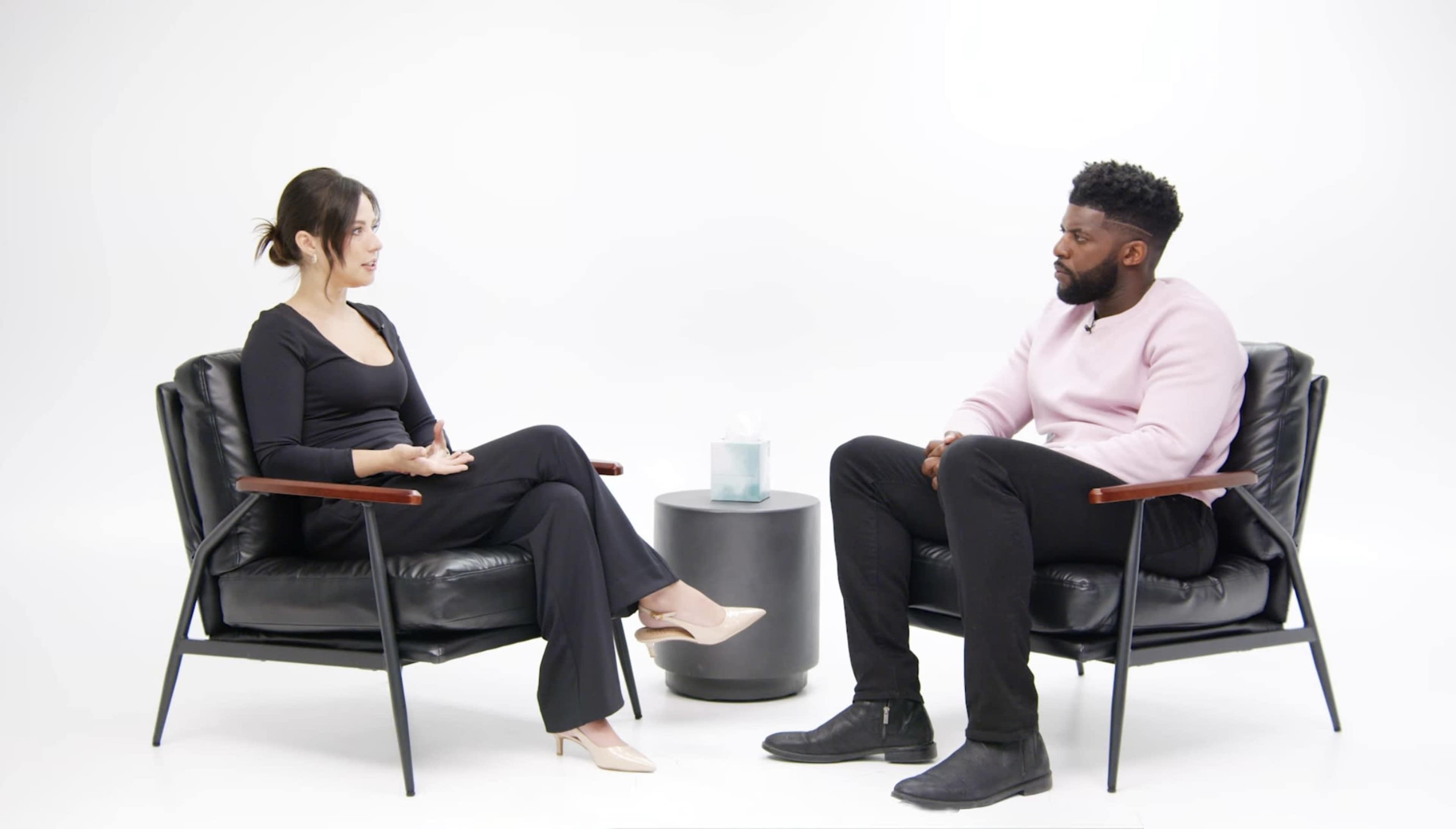 A woman and a man are seated in black armchairs facing each other, with a small round table between them.
