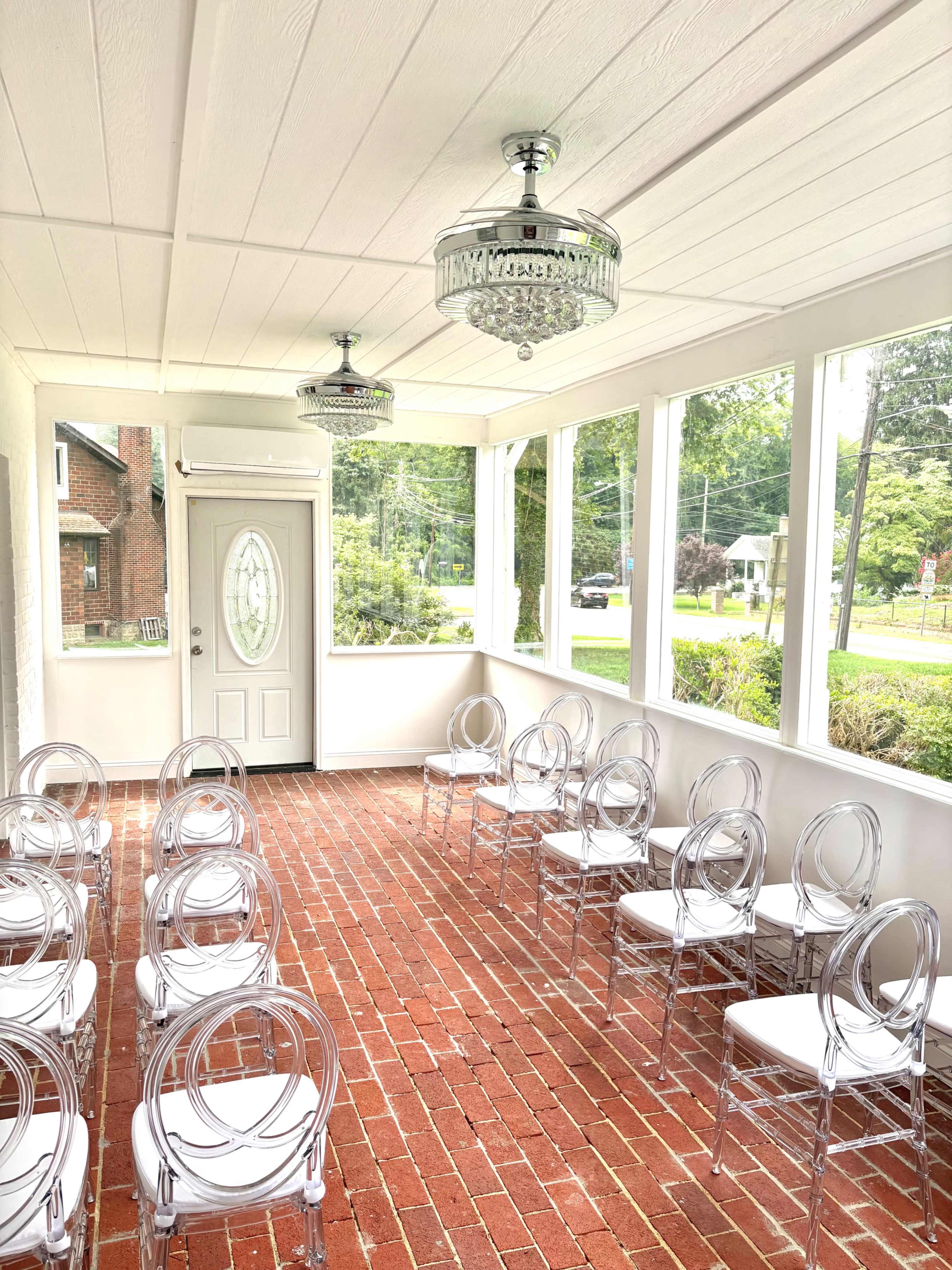 The image shows a bright and airy room with a brick floor, featuring several clear acrylic chairs arranged neatly in rows under two decorative chandeliers.