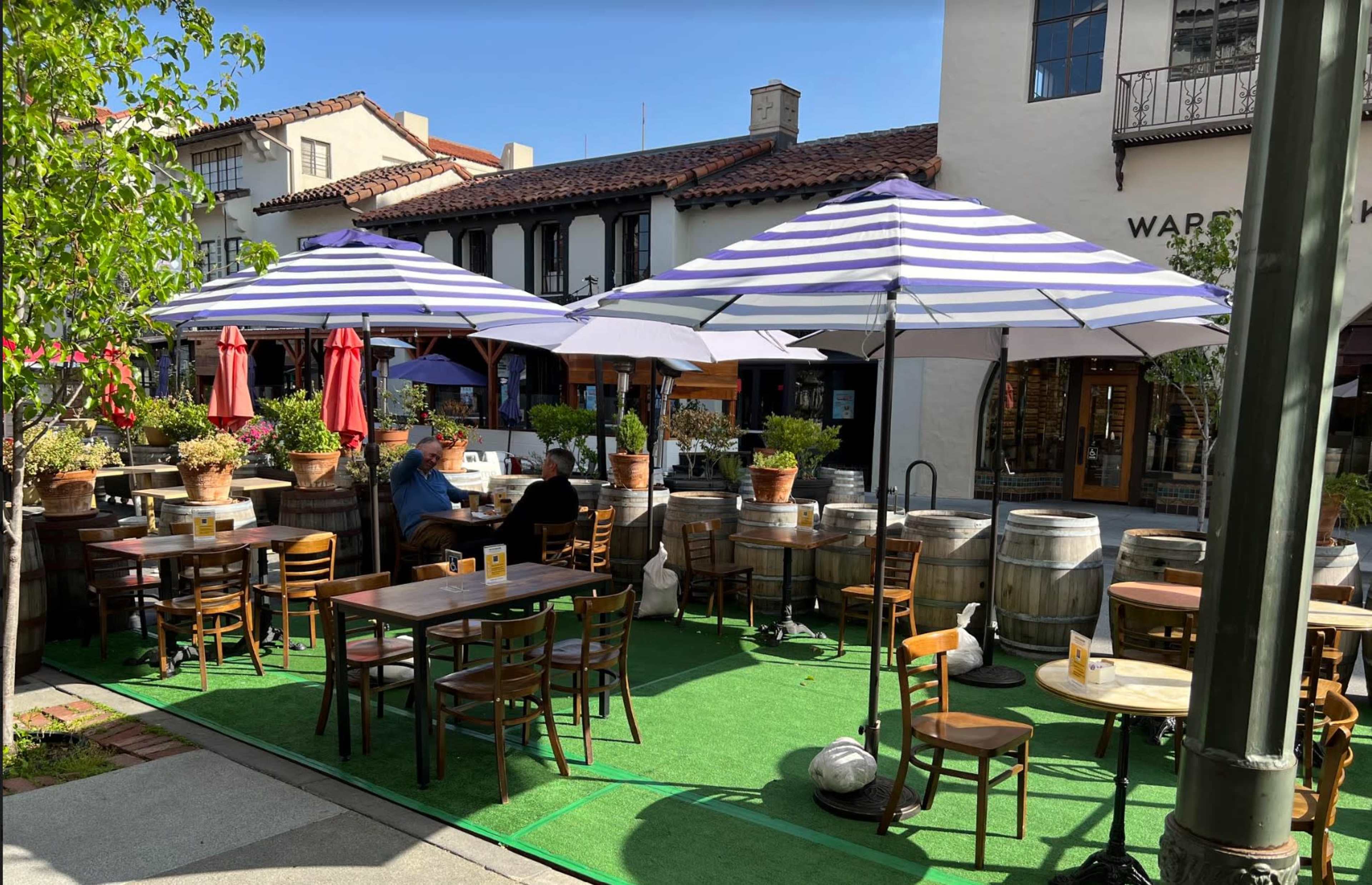 A shaded outdoor dining area with tables under purple and white striped umbrellas, surrounded by potted plants and barrels.