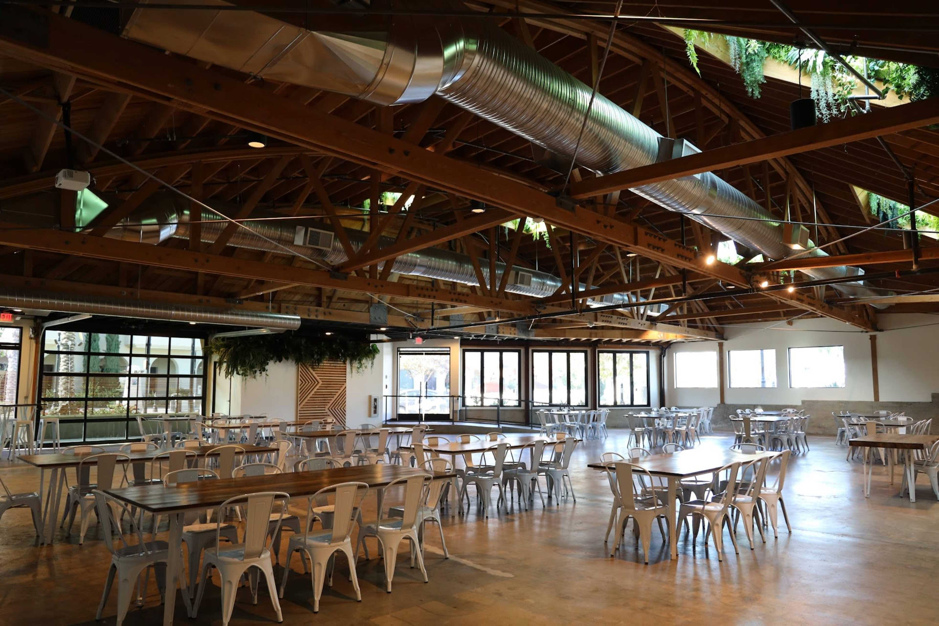 The image shows a spacious indoor event venue with numerous tables and chairs arranged neatly under a wooden ceiling.