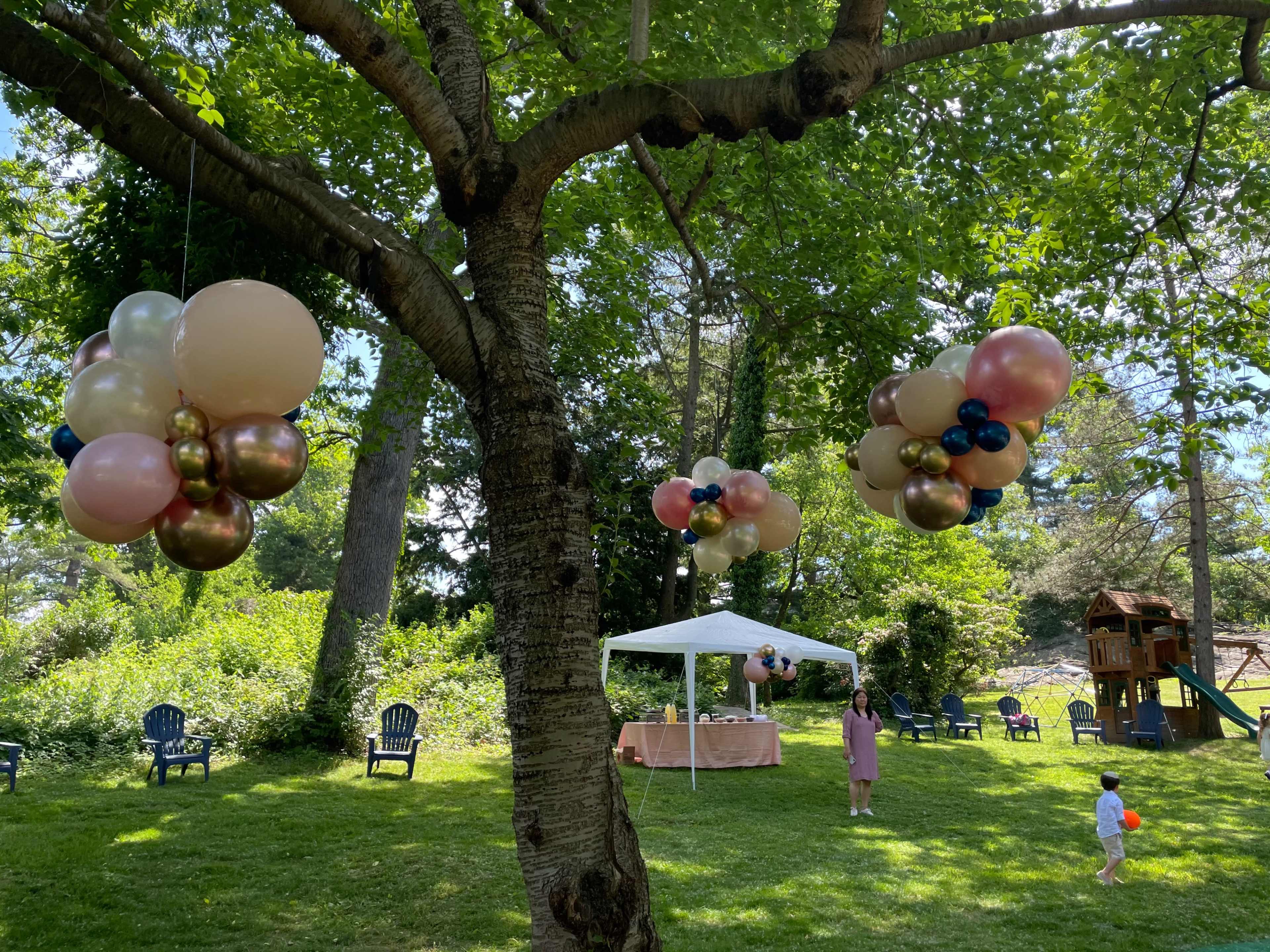A festive outdoor scene features decorative balloon arrangements hanging from a tree, a table set under a canopy, and a child playing nearby.