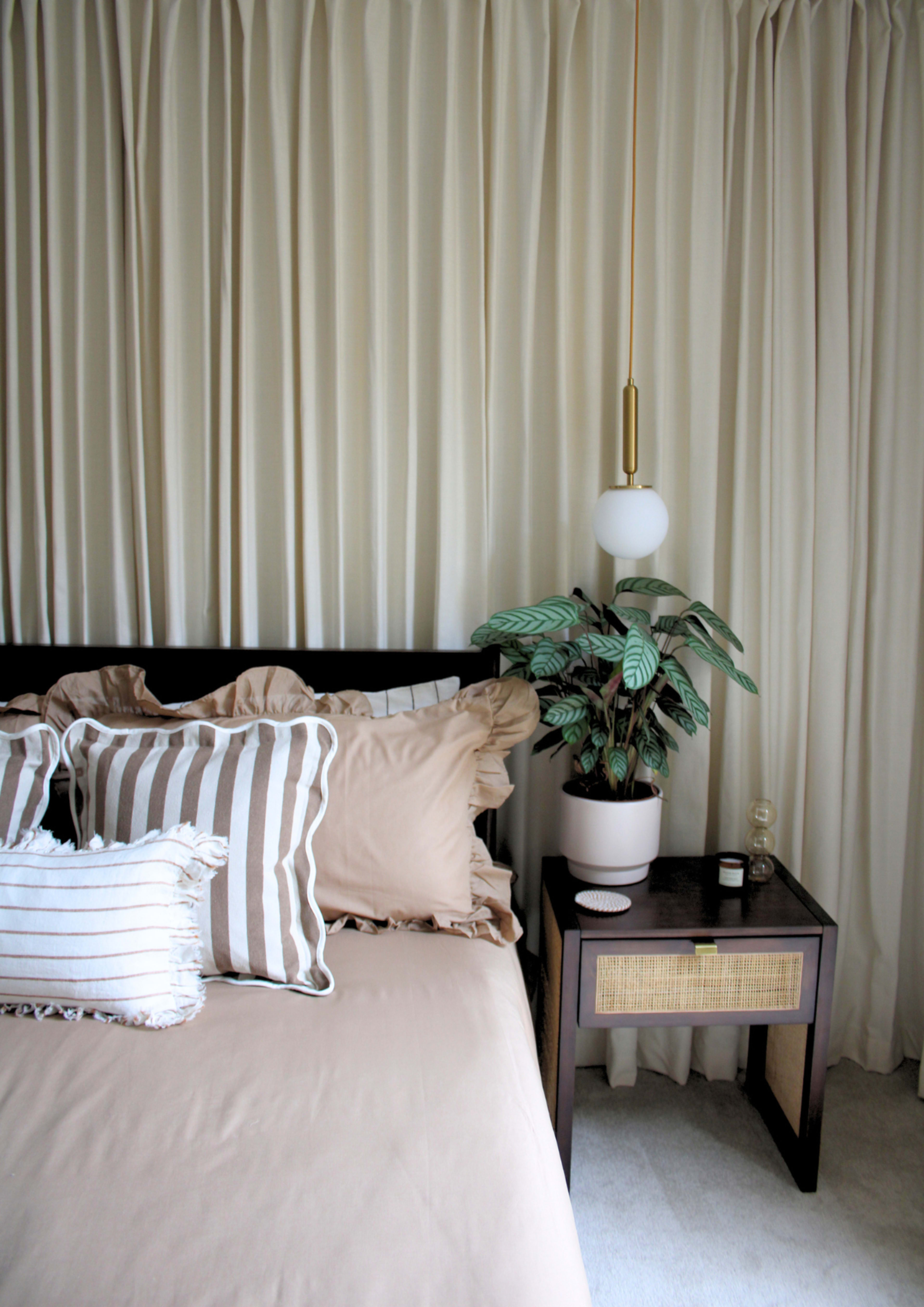 A bed with beige and striped pillows is positioned next to a dark wooden nightstand with a plant and decorative items, all set against a backdrop of cream-colored drapery.