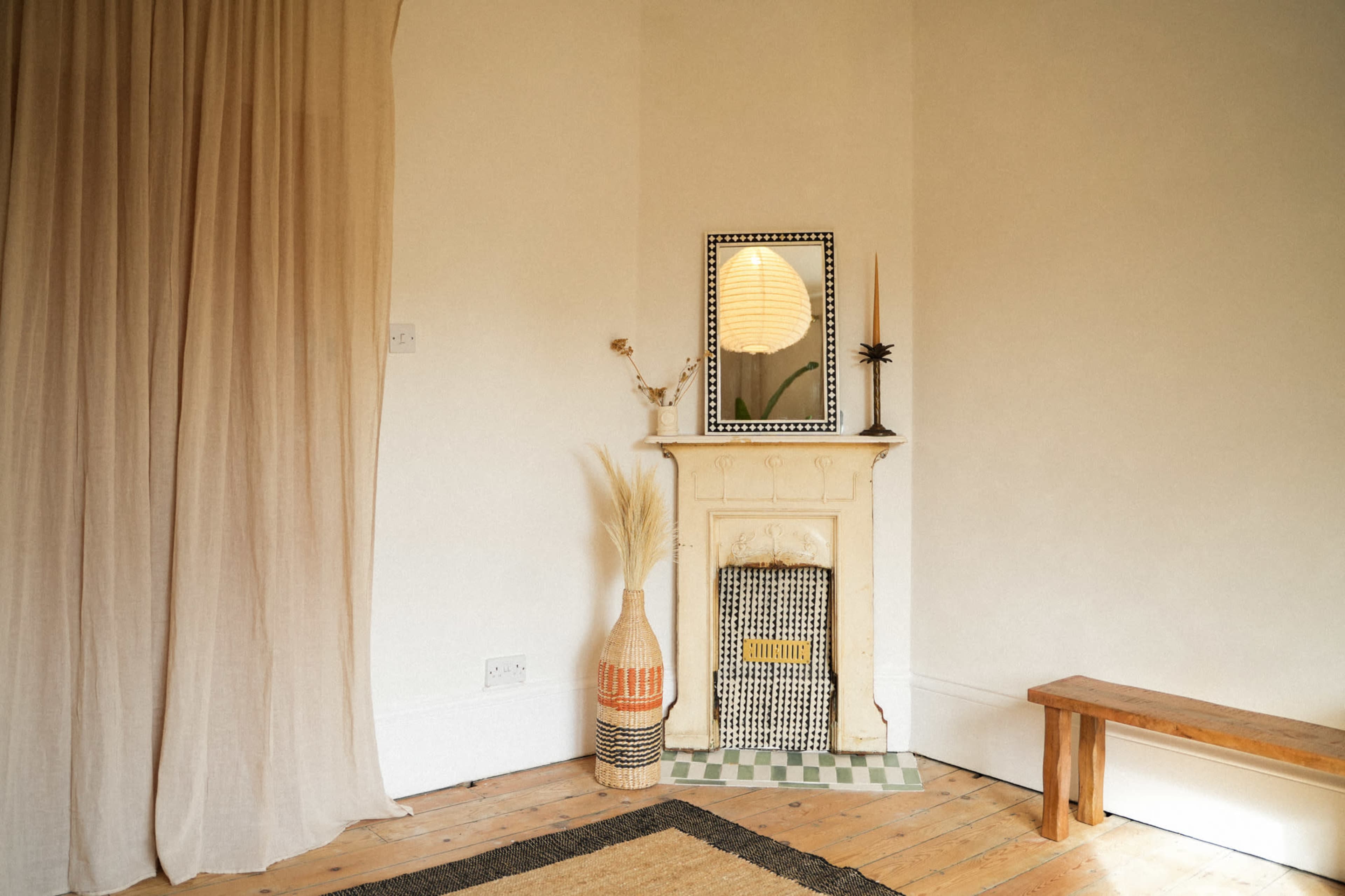 A small corner of a room features a vintage fireplace with a mirror above it, accompanied by a woven basket and a wooden bench.