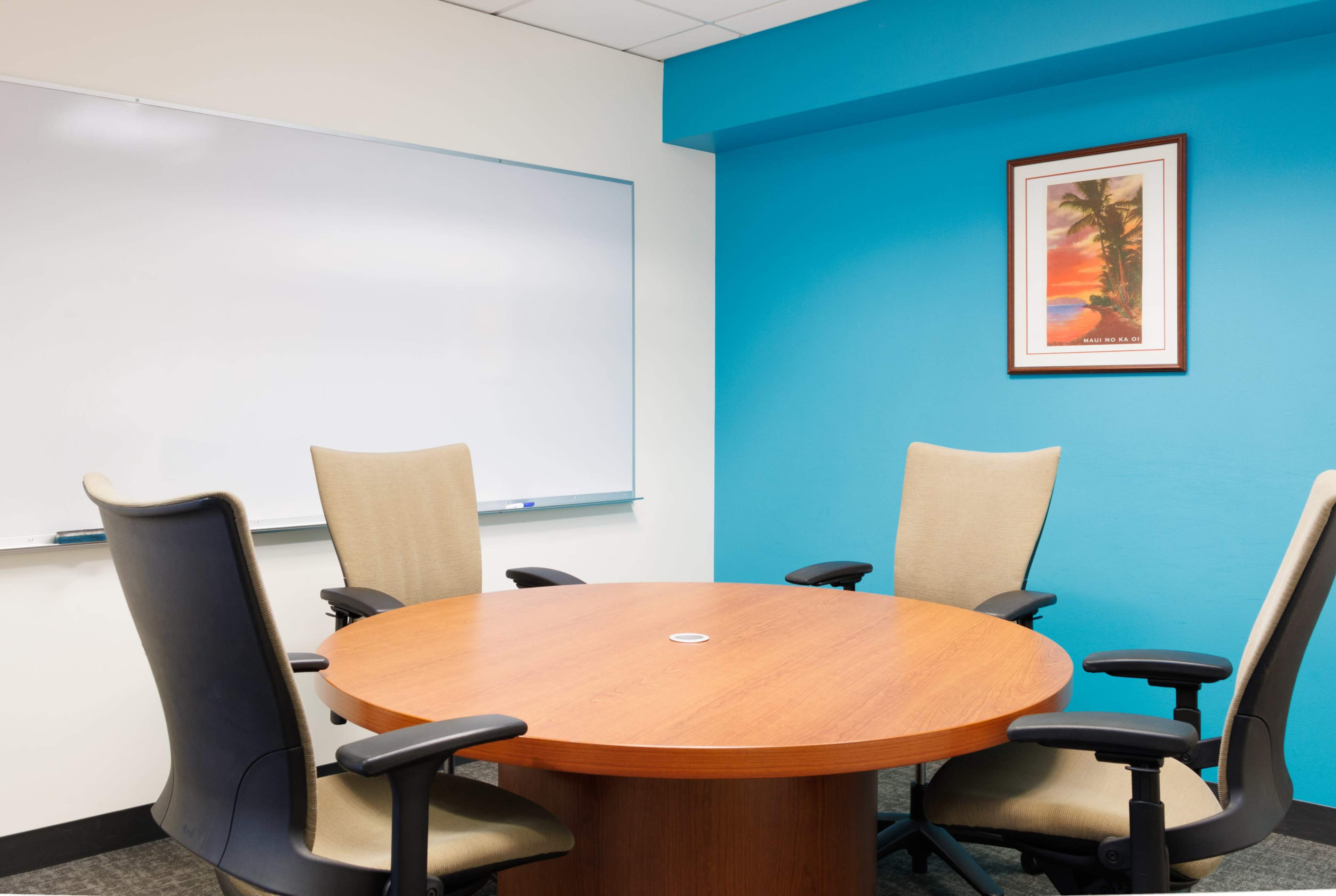 A round wooden conference table surrounded by four chairs in a brightly colored meeting room with a whiteboard and a framed picture on the wall.