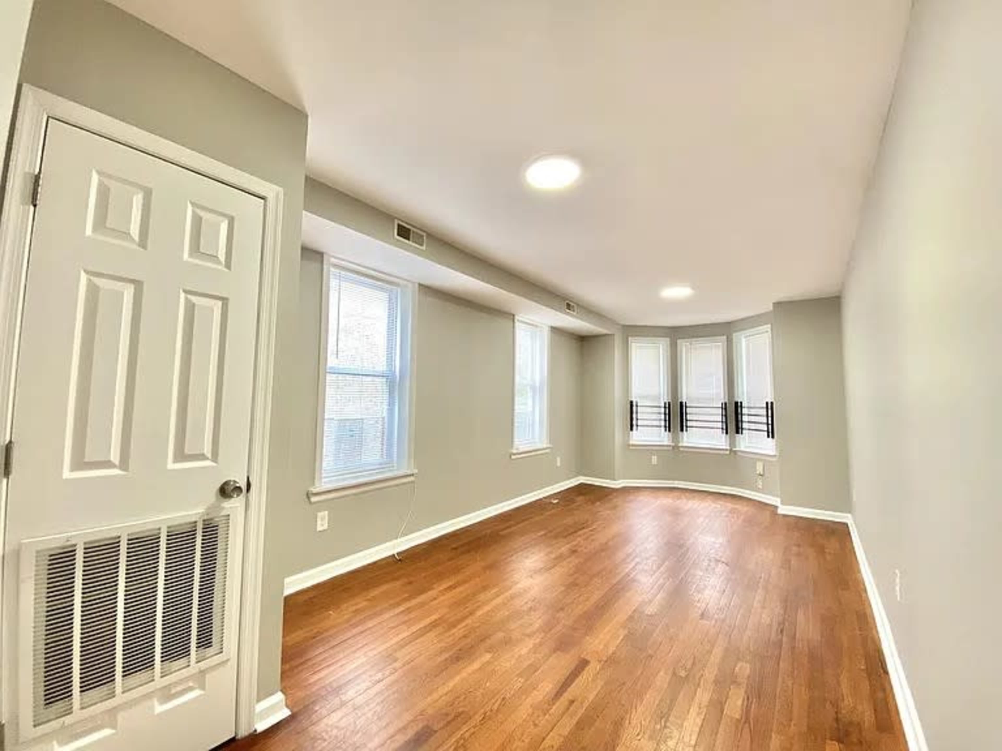 The image shows an empty room with hardwood floors, large windows, and a light gray painted wall.