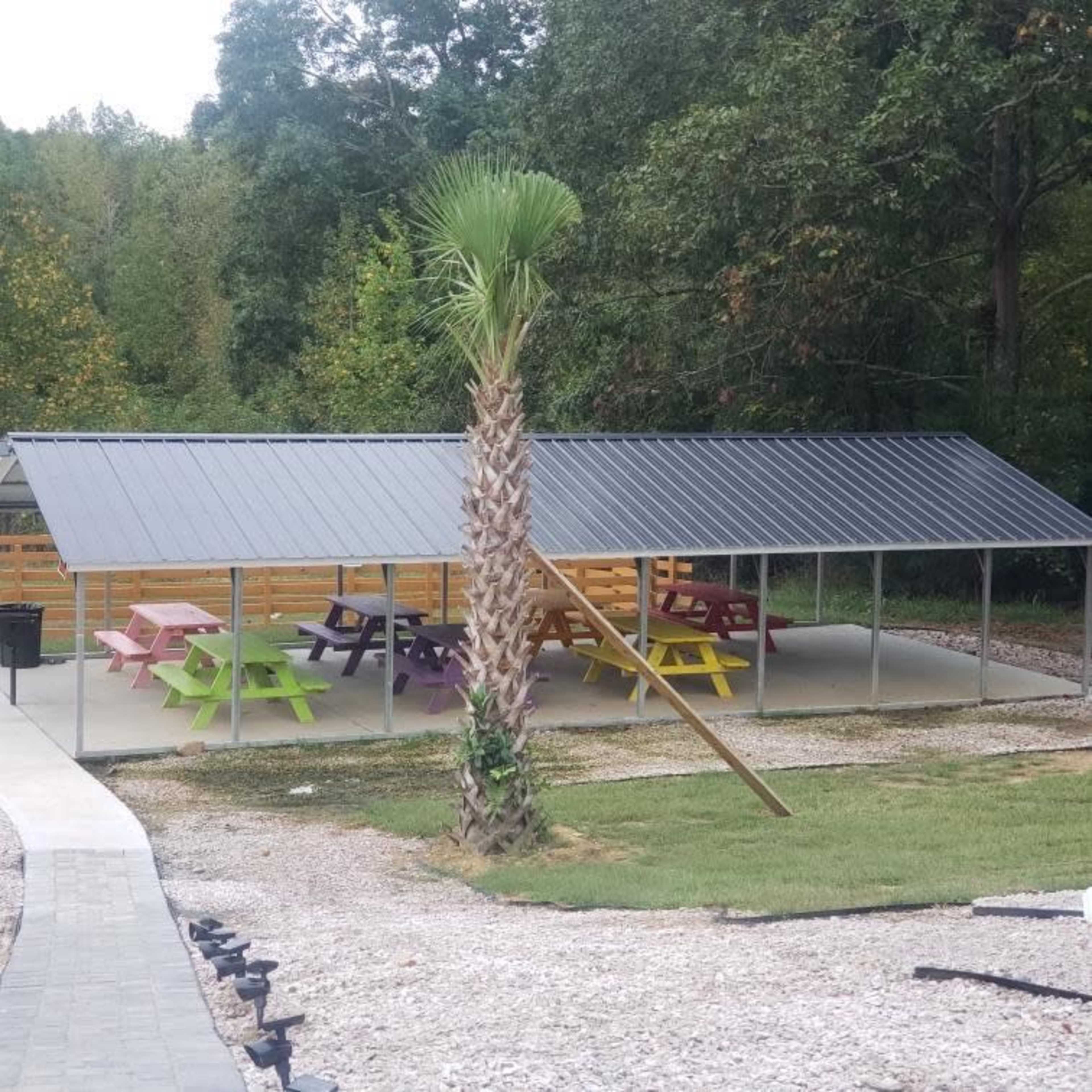 The image shows a metal-roofed pavilion with colorful picnic tables underneath, located near a palm tree and surrounded by a gravel area.