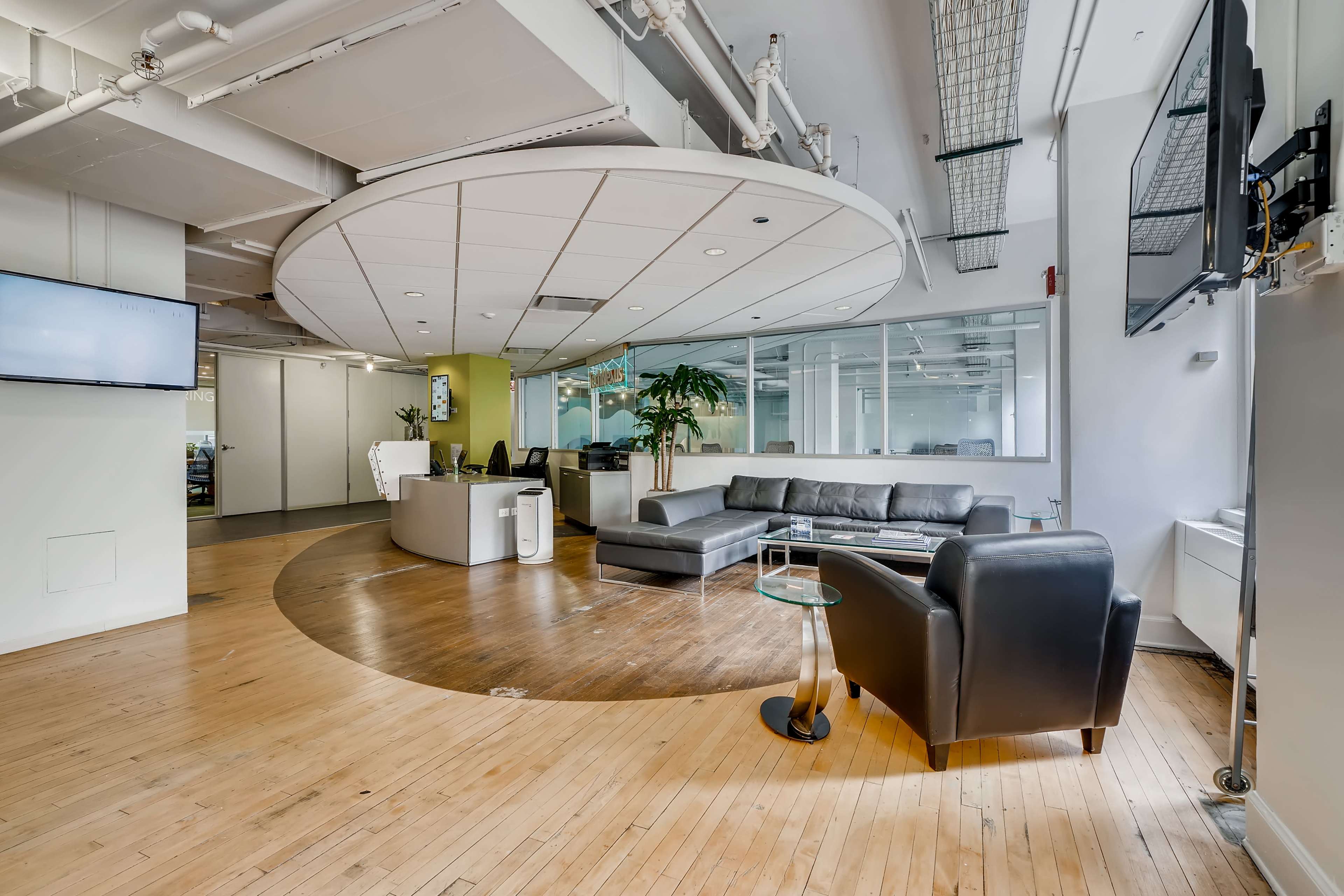 The image shows a modern office reception area with a seating arrangement, including a black sofa and armchair, along with a circular wood floor design and large windows illuminating the space.