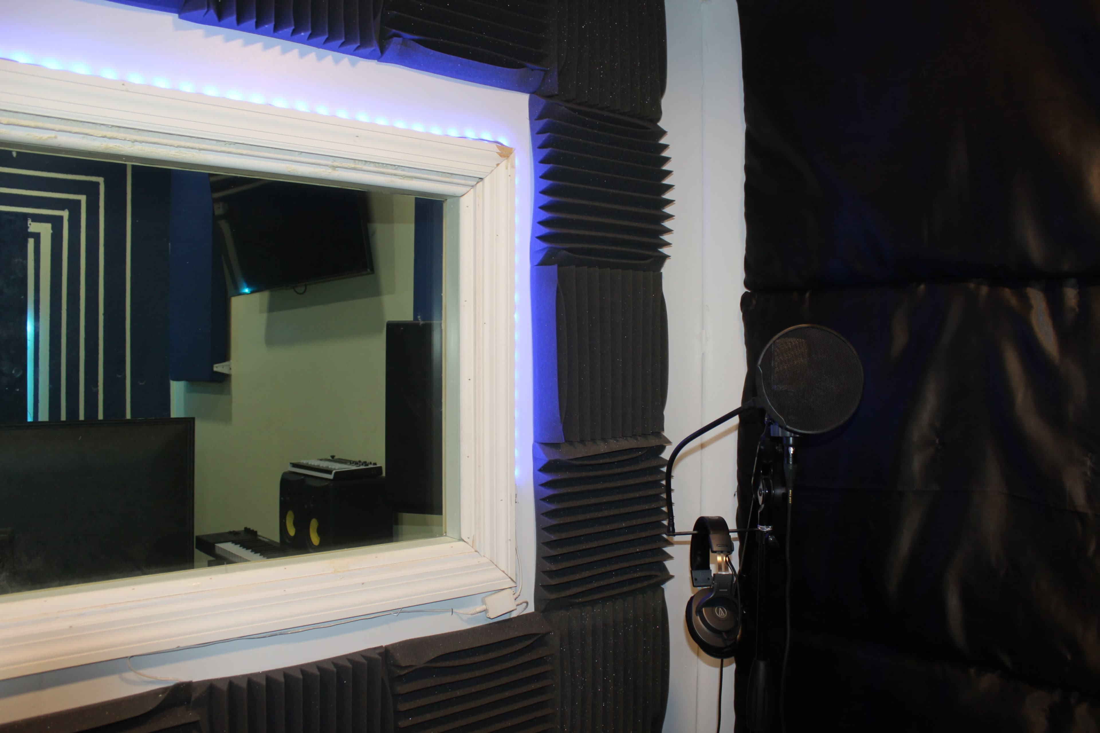The image shows a recording studio booth with soundproofing panels, a microphone on a stand, and headphones hanging nearby.