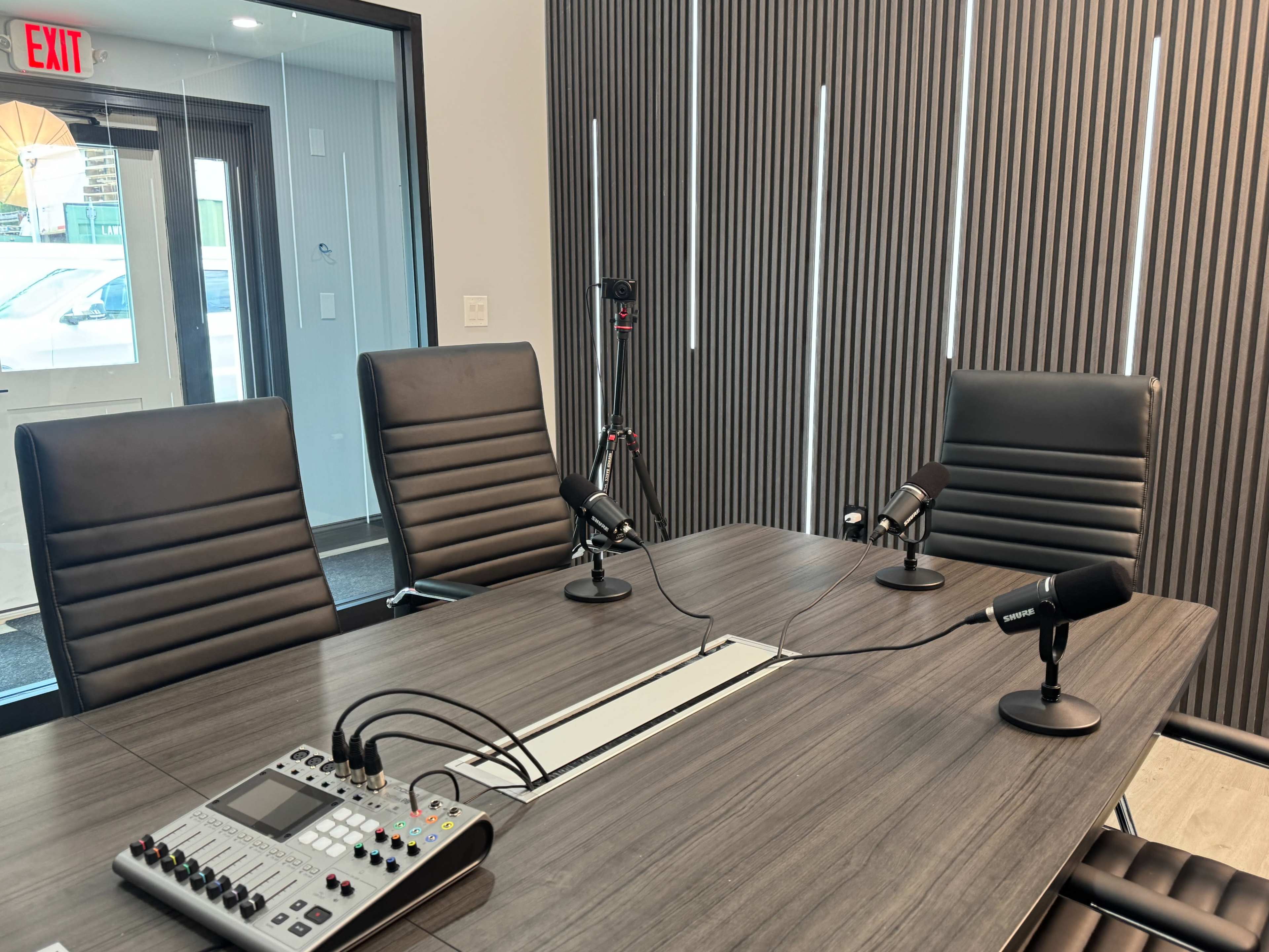 A modern conference room features a rectangular table with four black chairs, microphones, and audio equipment, set against a backdrop of vertical wood paneling.