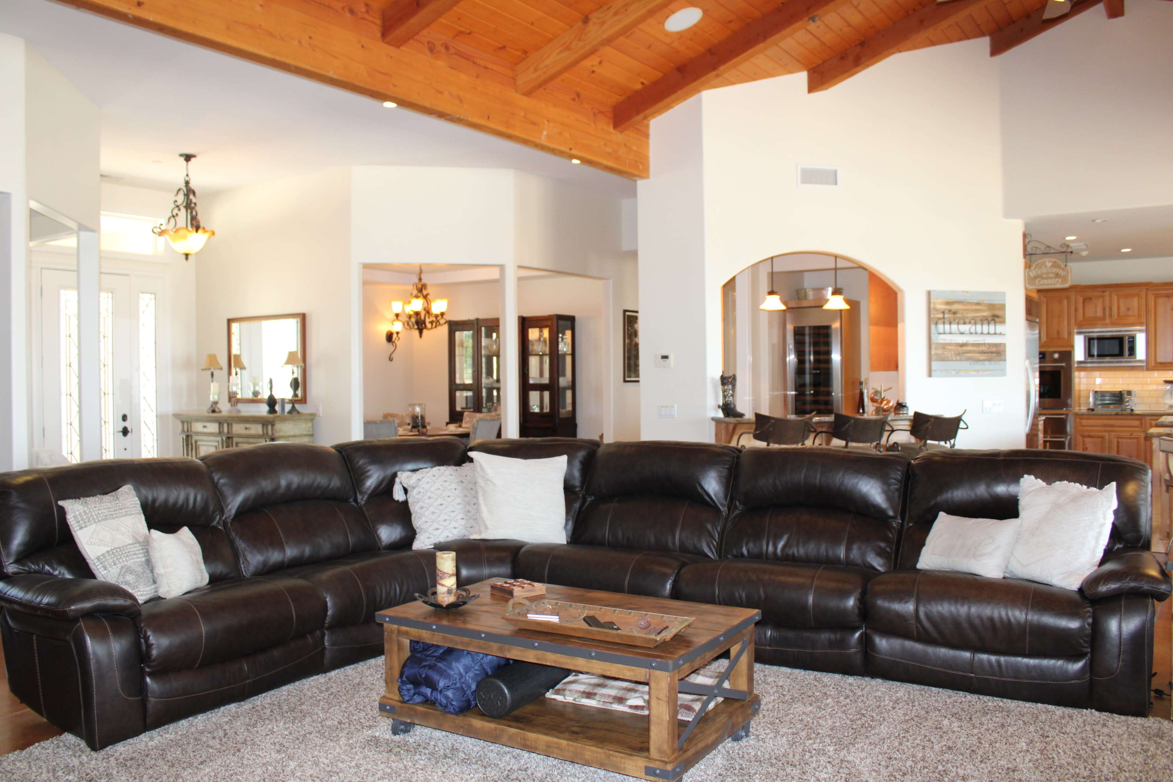 The image shows a spacious living room with a dark leather sectional couch, a wooden coffee table, and a view of a kitchen and dining area in the background.