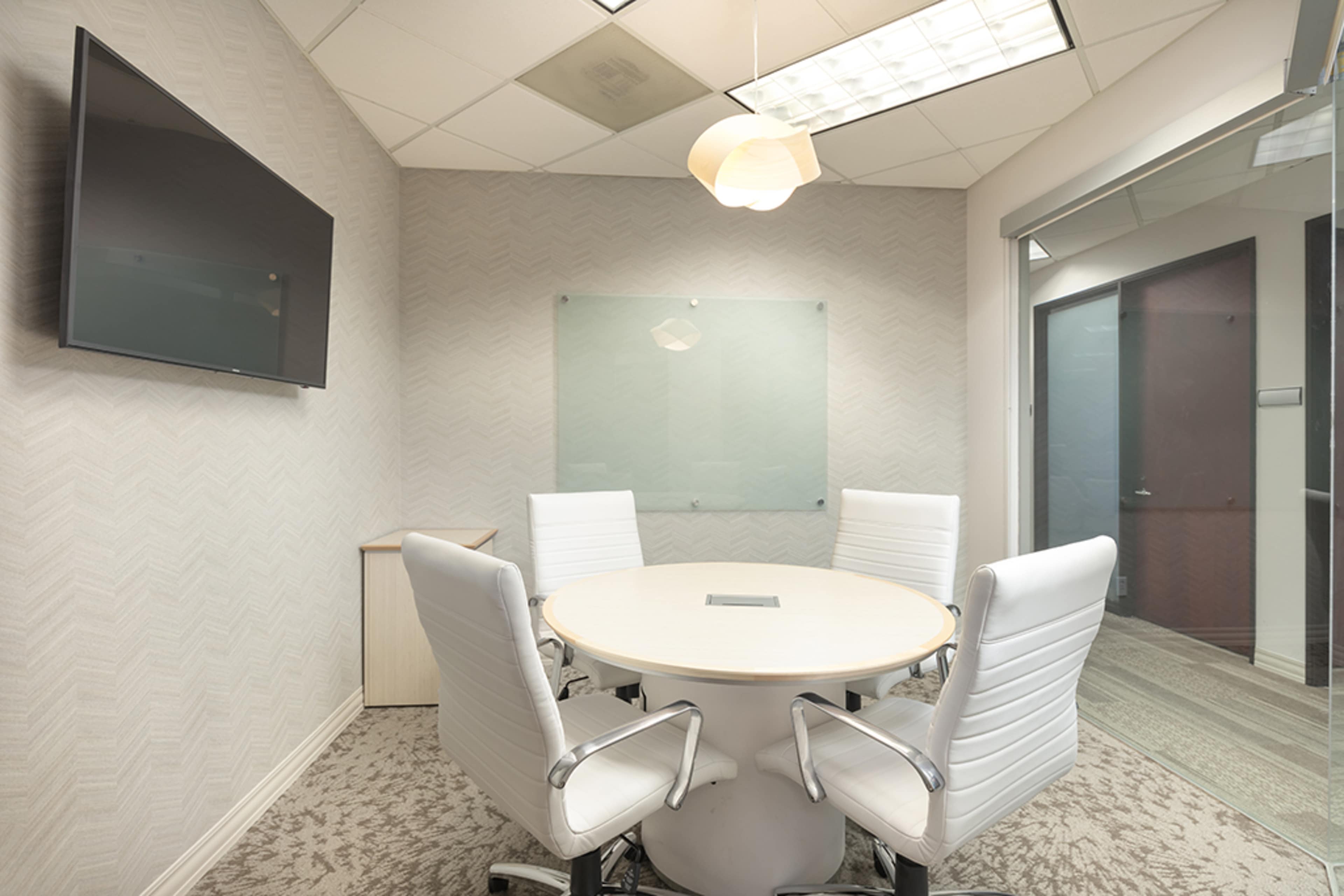 The image shows a modern conference room with a round table, four white chairs, a wall-mounted TV, and a glass partition.