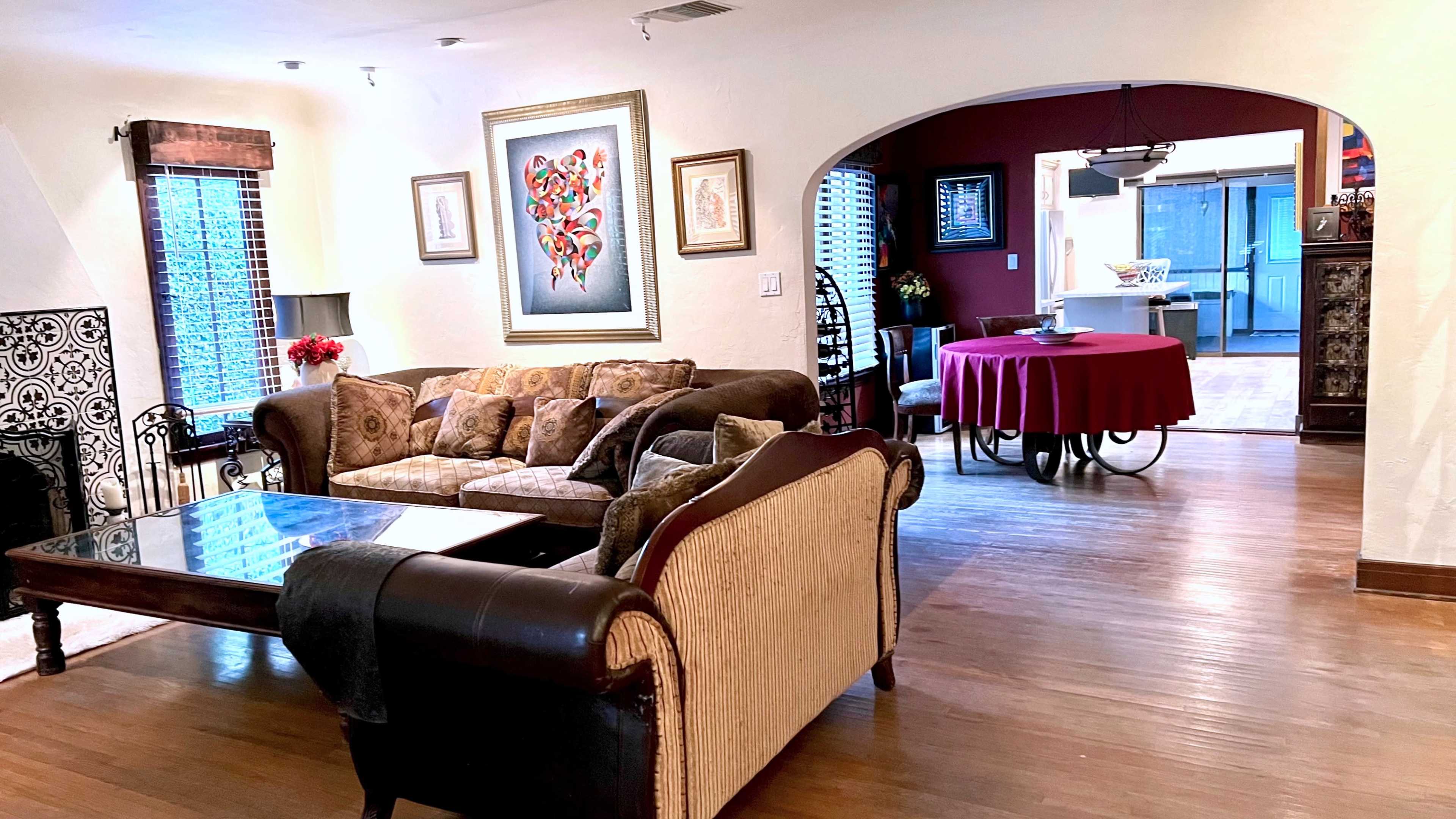 A living room with a couch, armchair, and coffee table, leading into a dining area with a round table covered by a red tablecloth.