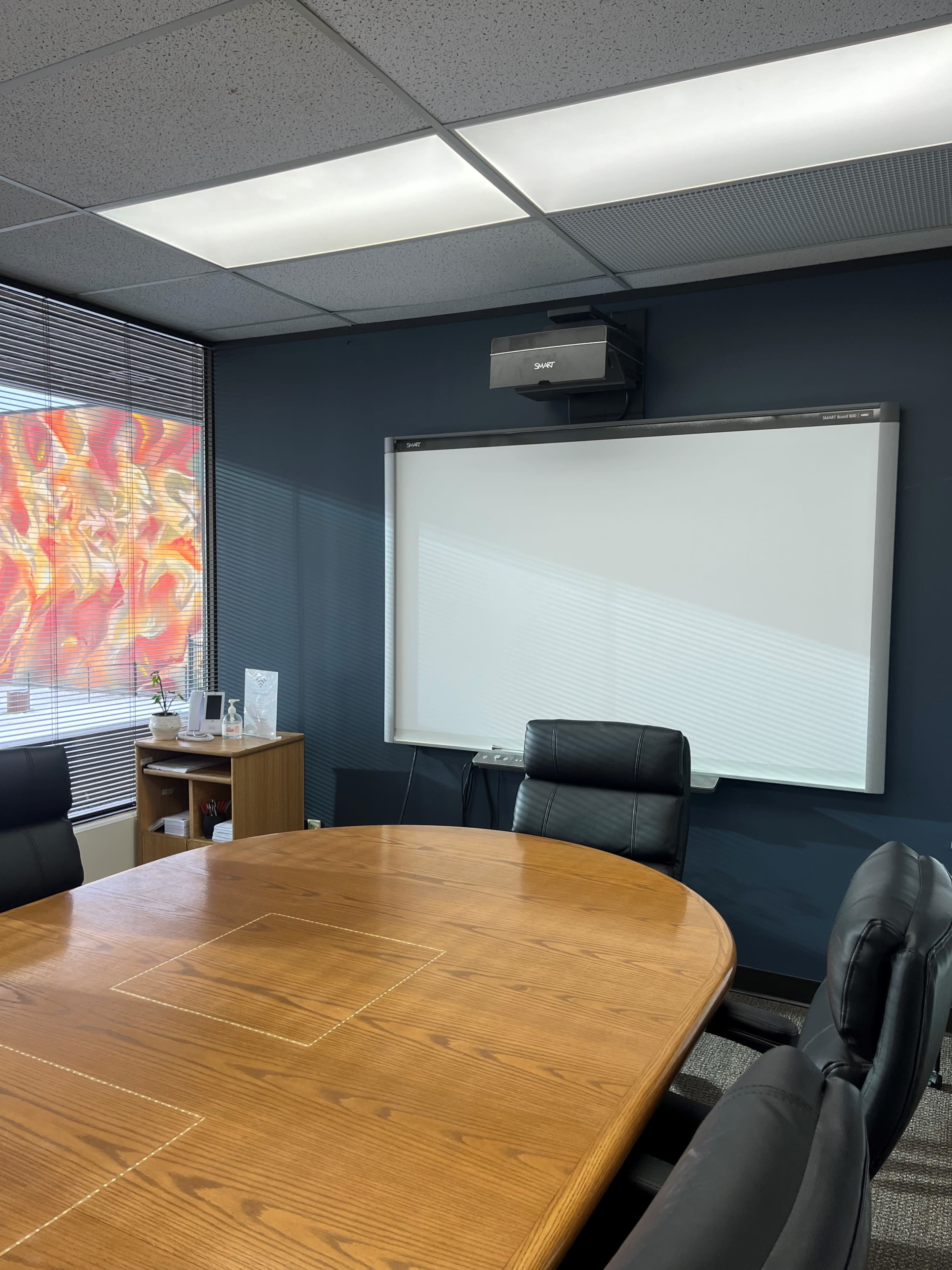 The image shows a conference room with a large wooden table, black leather chairs, a wall-mounted projector, and a blank white projection screen.