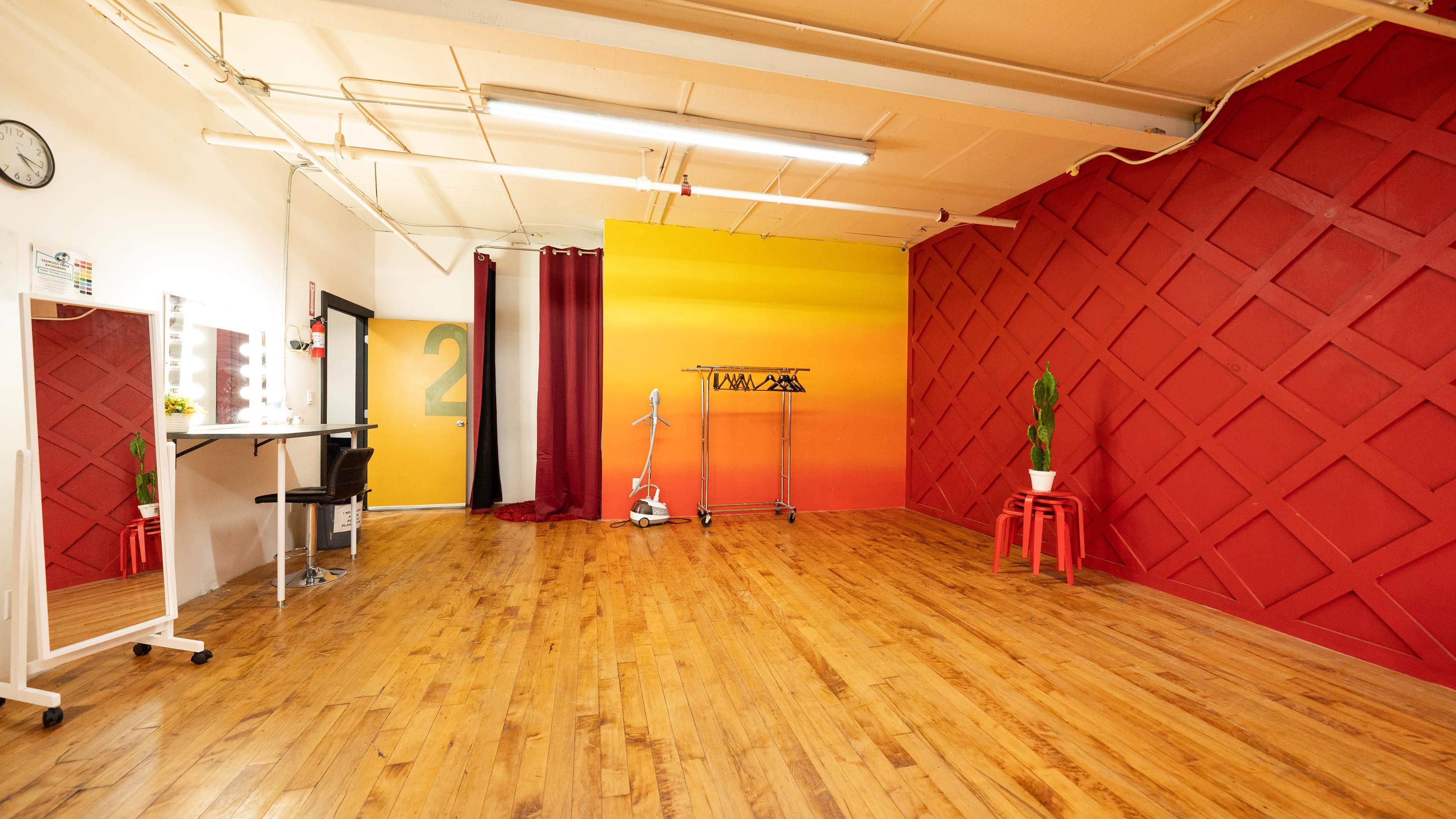 The image shows a brightly lit studio space featuring wooden floors, a mirrored vanity, a clothing rack, and a painted wall with a gradient from yellow to orange, alongside a red accent wall.