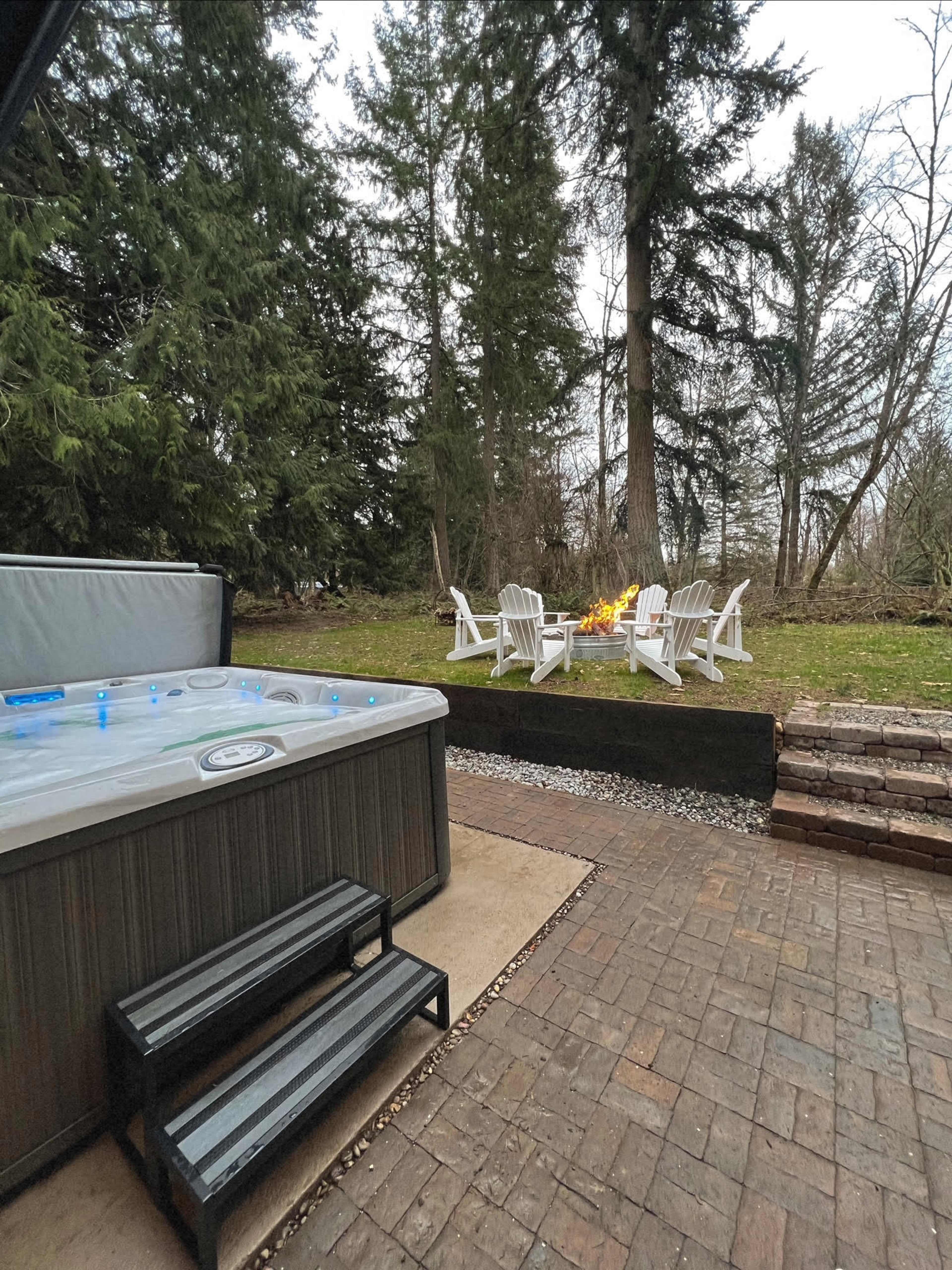 A hot tub beside a patio area with Adirondack chairs arranged around a fire pit, set against a backdrop of trees.