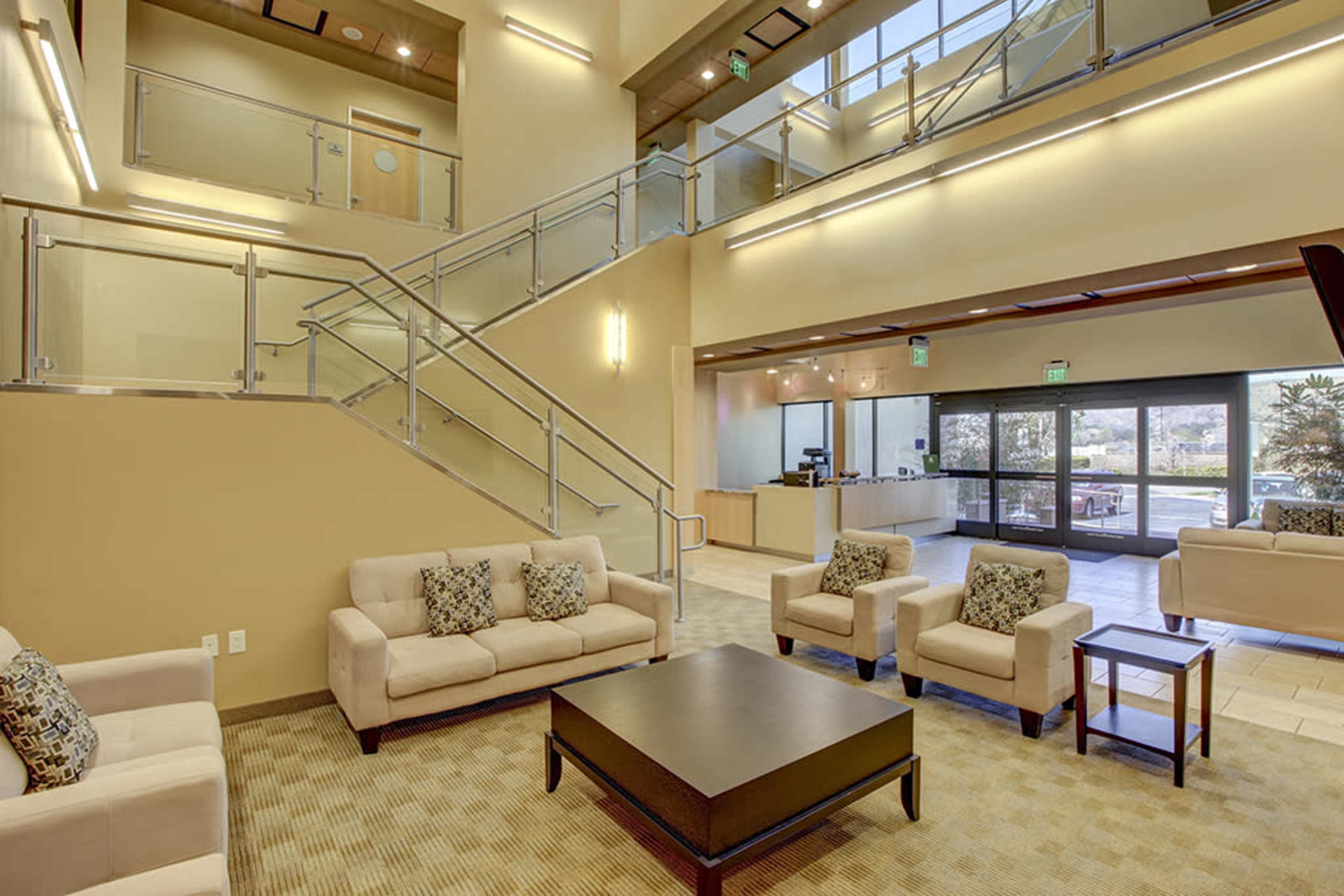 A modern lobby area featuring beige furniture, a large coffee table, and a staircase with glass railing.