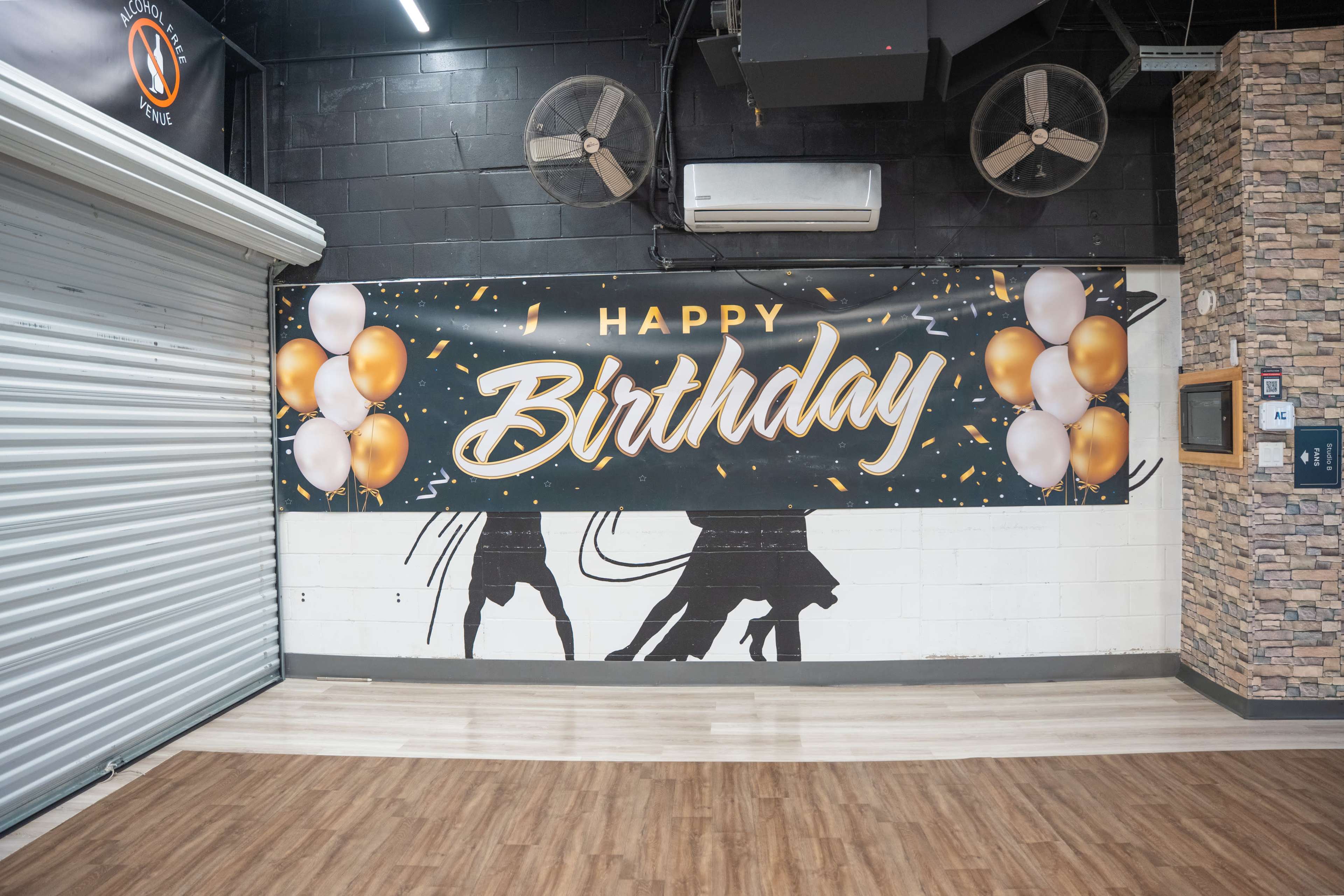 A large "Happy Birthday" banner with balloons is mounted on a wall inside a venue, with a shuttered door visible nearby.