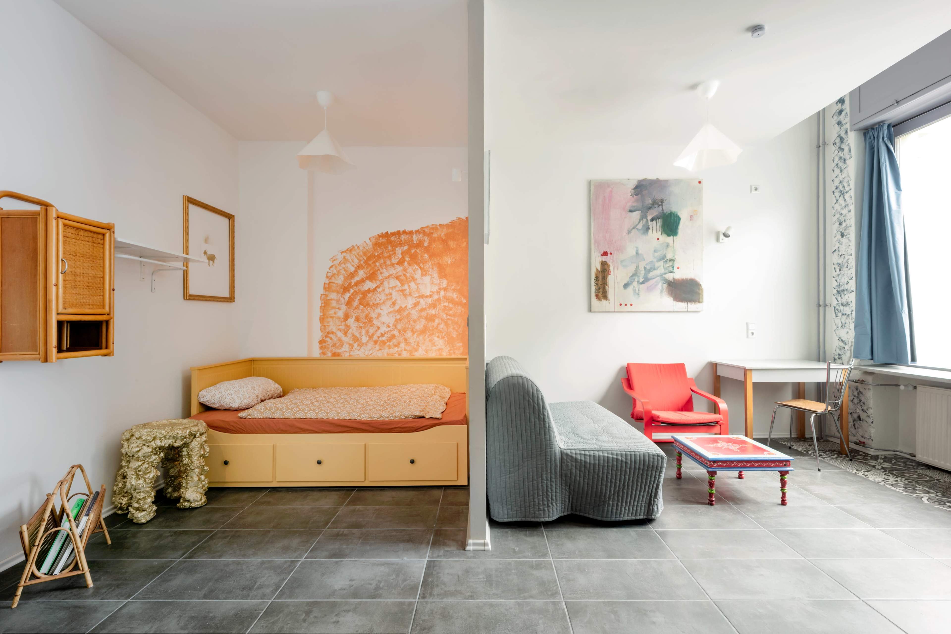 The image shows a modern studio room divided into sleeping and living areas, featuring a bed, a small table, a chair, and decorative wall art.
