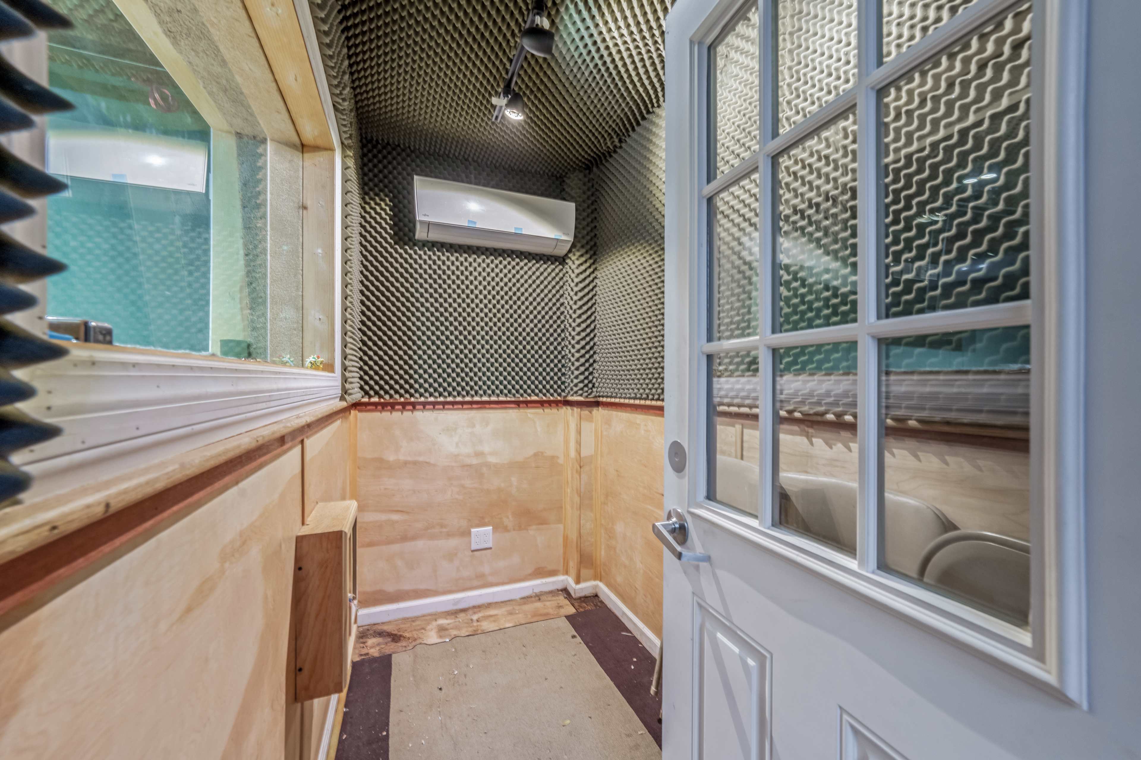 The image shows a small soundproof room with acoustic foam panels covering the walls, featuring a glass window and a white door.