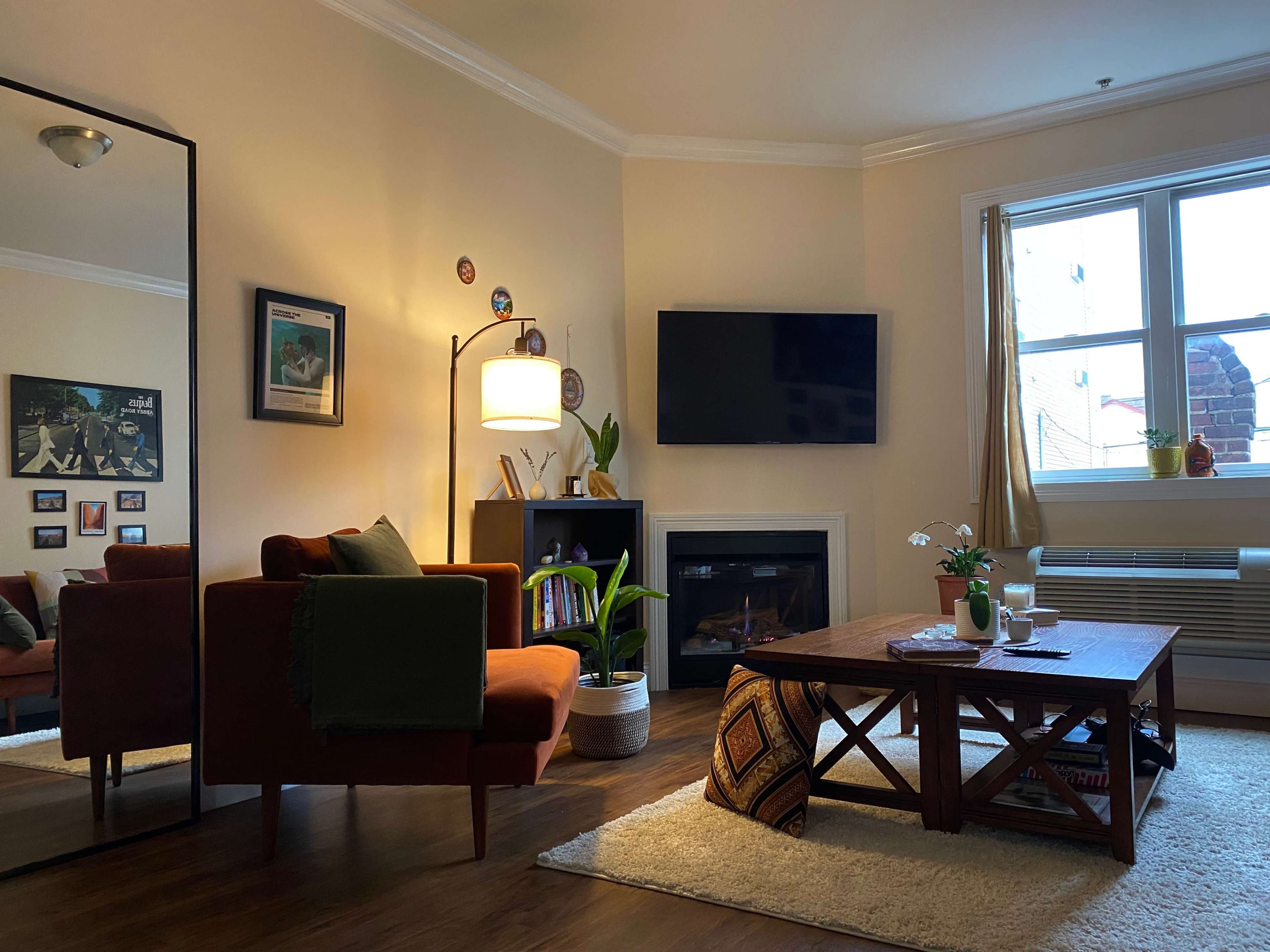 A cozy living room featuring a brown couch, a wooden coffee table, a television mounted on the wall, and a fireplace, with plants and decorative elements present.