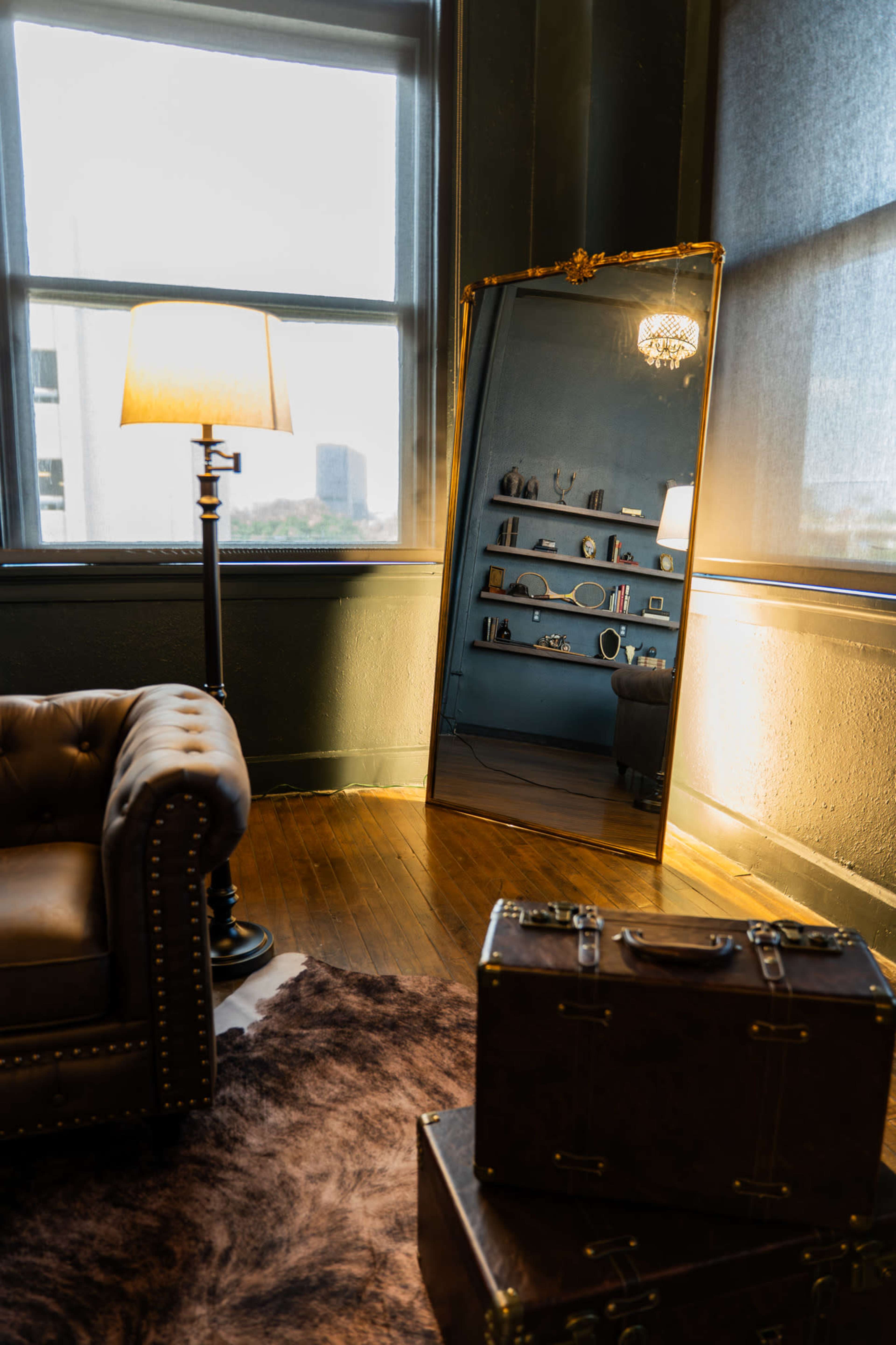 The image shows a cozy room featuring a tufted leather sofa, a floor lamp, a large ornate mirror, and a shelf displaying various small objects.