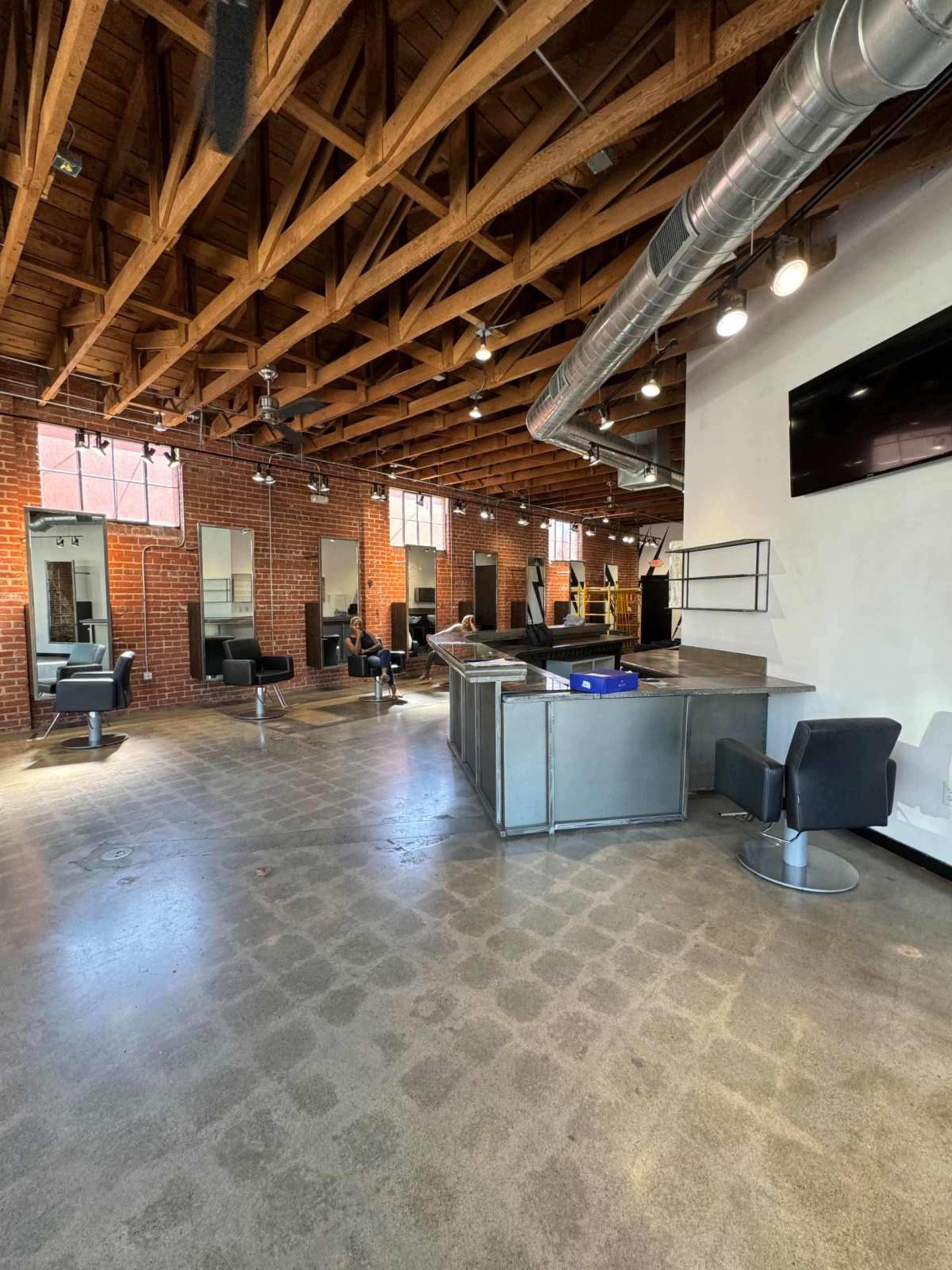 The image shows a modern salon space with a polished concrete floor, exposed brick walls, and several styling stations equipped with mirrors and chairs under a wooden ceiling.