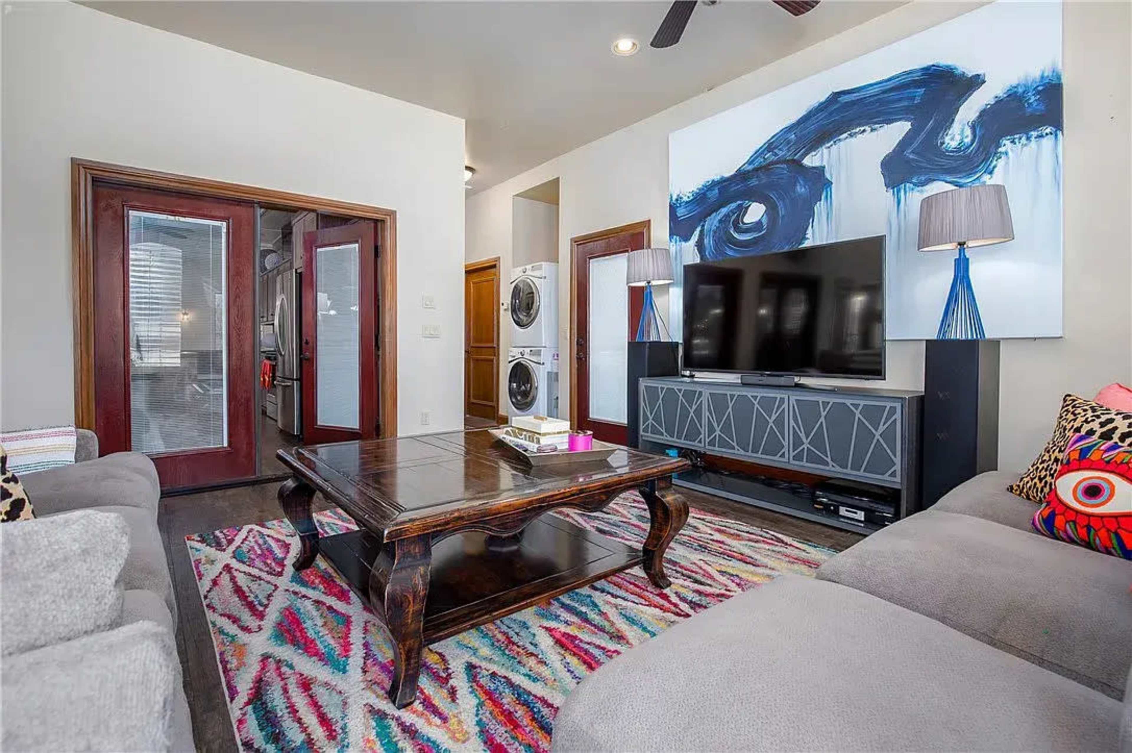 The living room features a coffee table in front of a large television, flanked by two modern lamps, with a colorful rug beneath and a washing machine visible in the background.