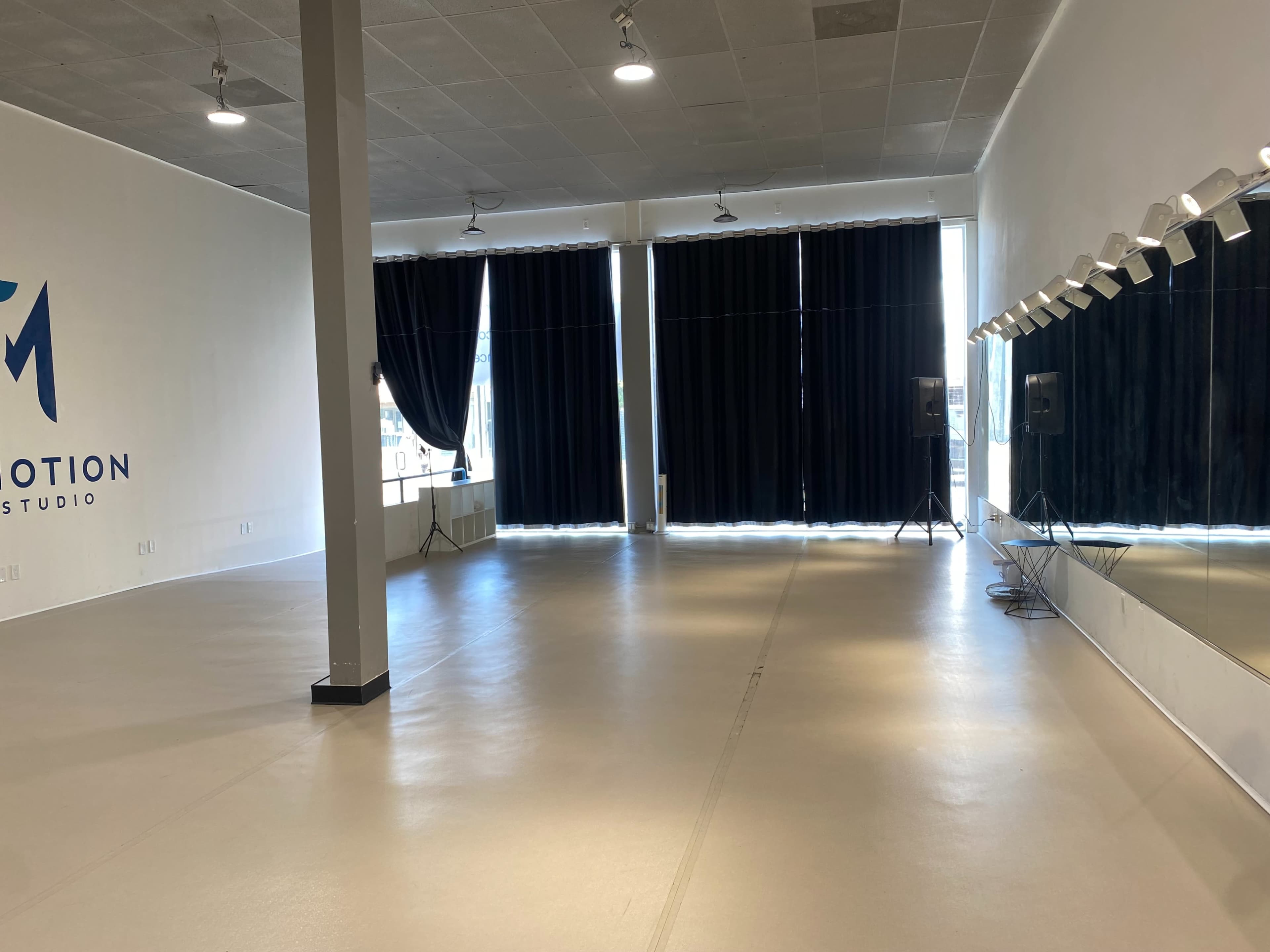 A spacious dance studio with large windows covered by black curtains, a mirror along one wall, and sound equipment set up in the corners.