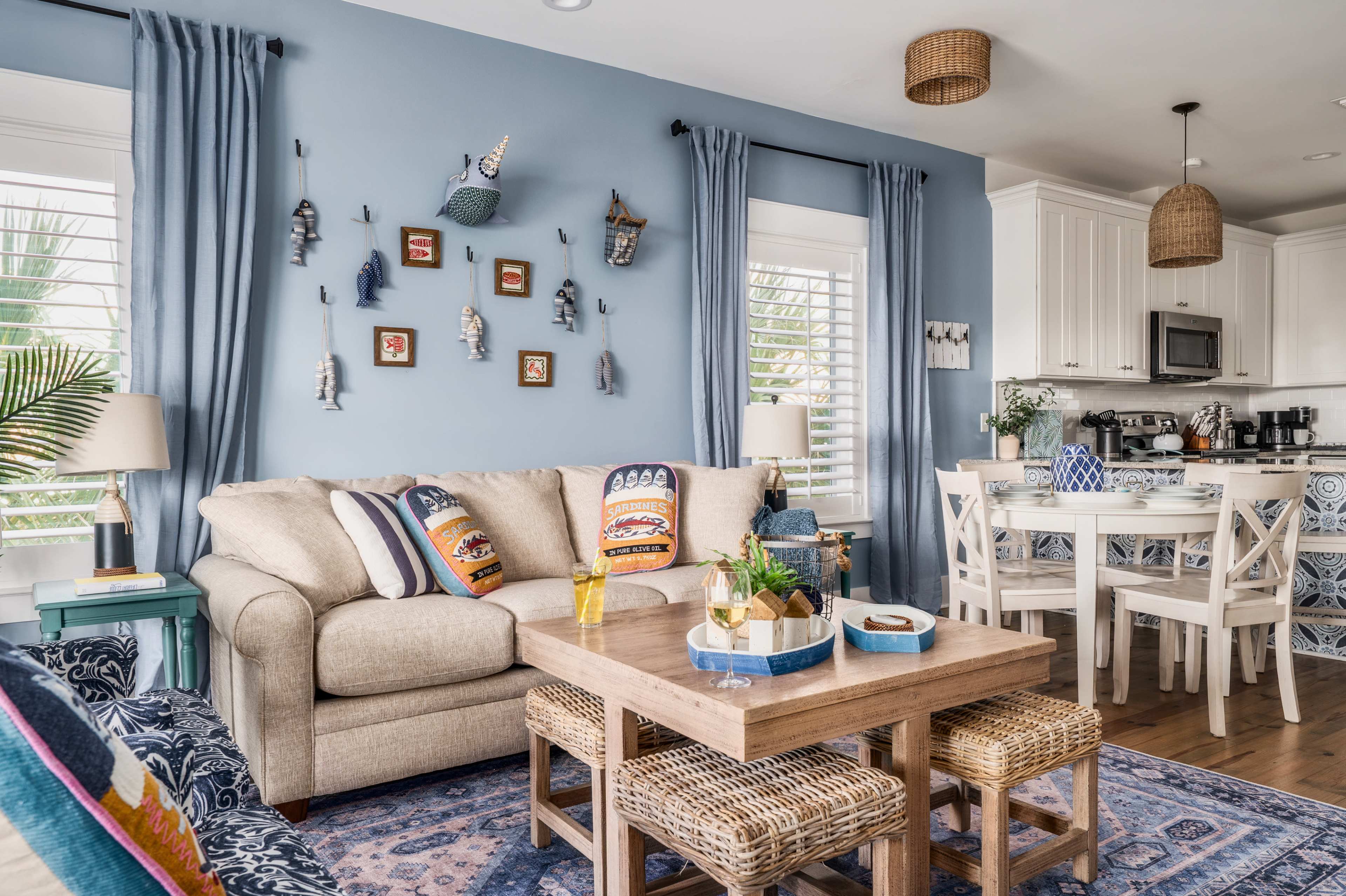 The image shows a brightly lit living room with a beige sofa, decorative pillows, a wooden coffee table, and a kitchen area in the background with white cabinetry.