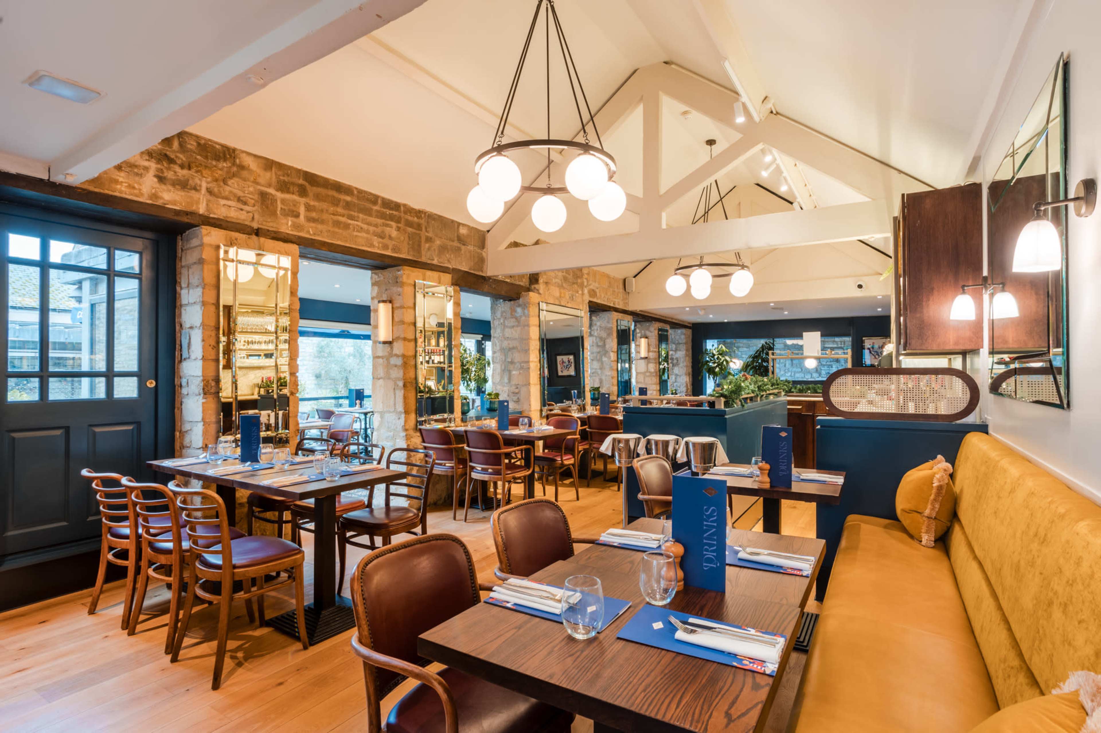 The image shows a modern restaurant interior with wooden floors, a mix of tables and seating, and large windows letting in natural light.
