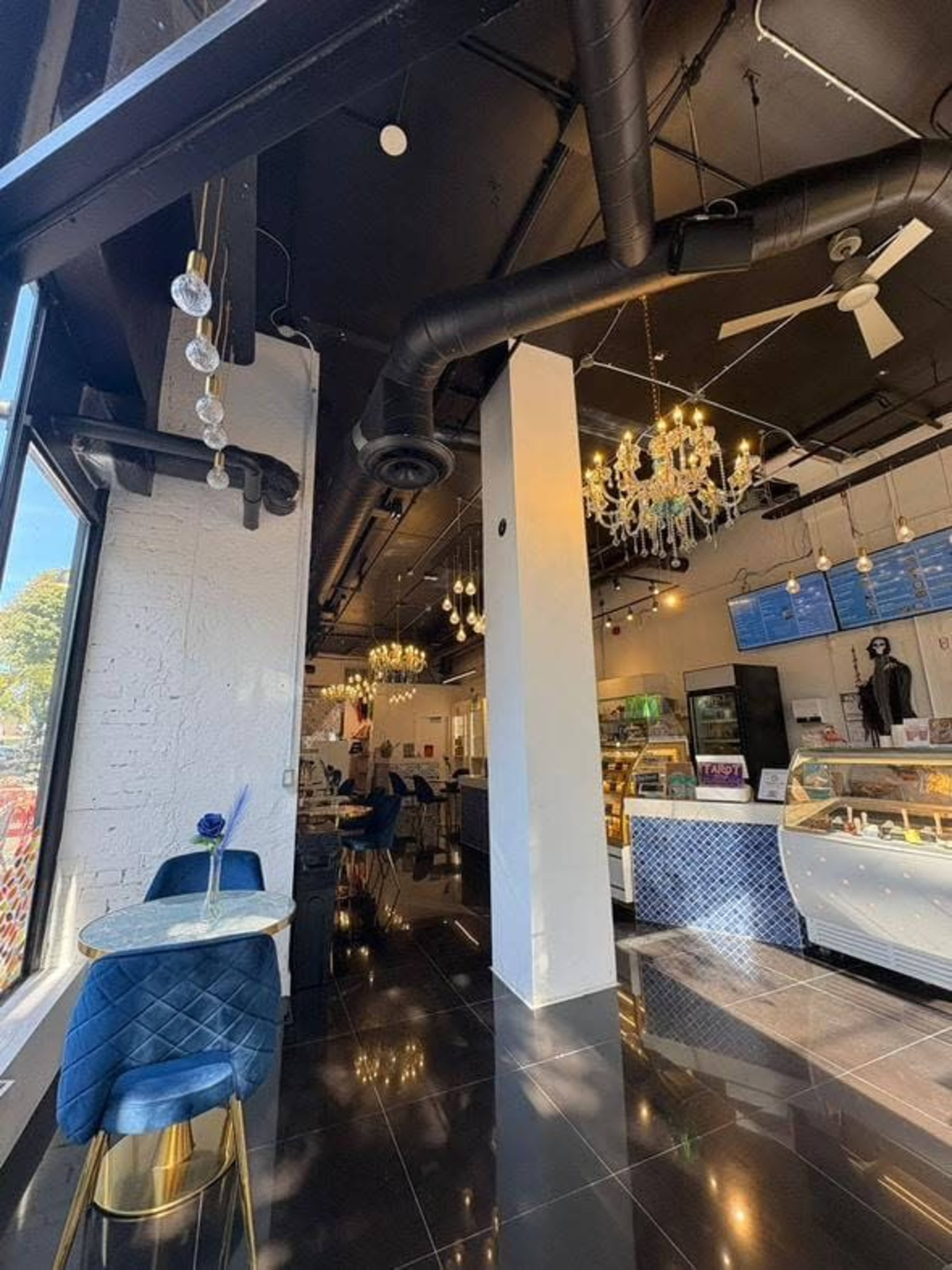 The image shows a modern café interior featuring dark tile flooring, elegant chandeliers, and displays of pastries in glass cases.