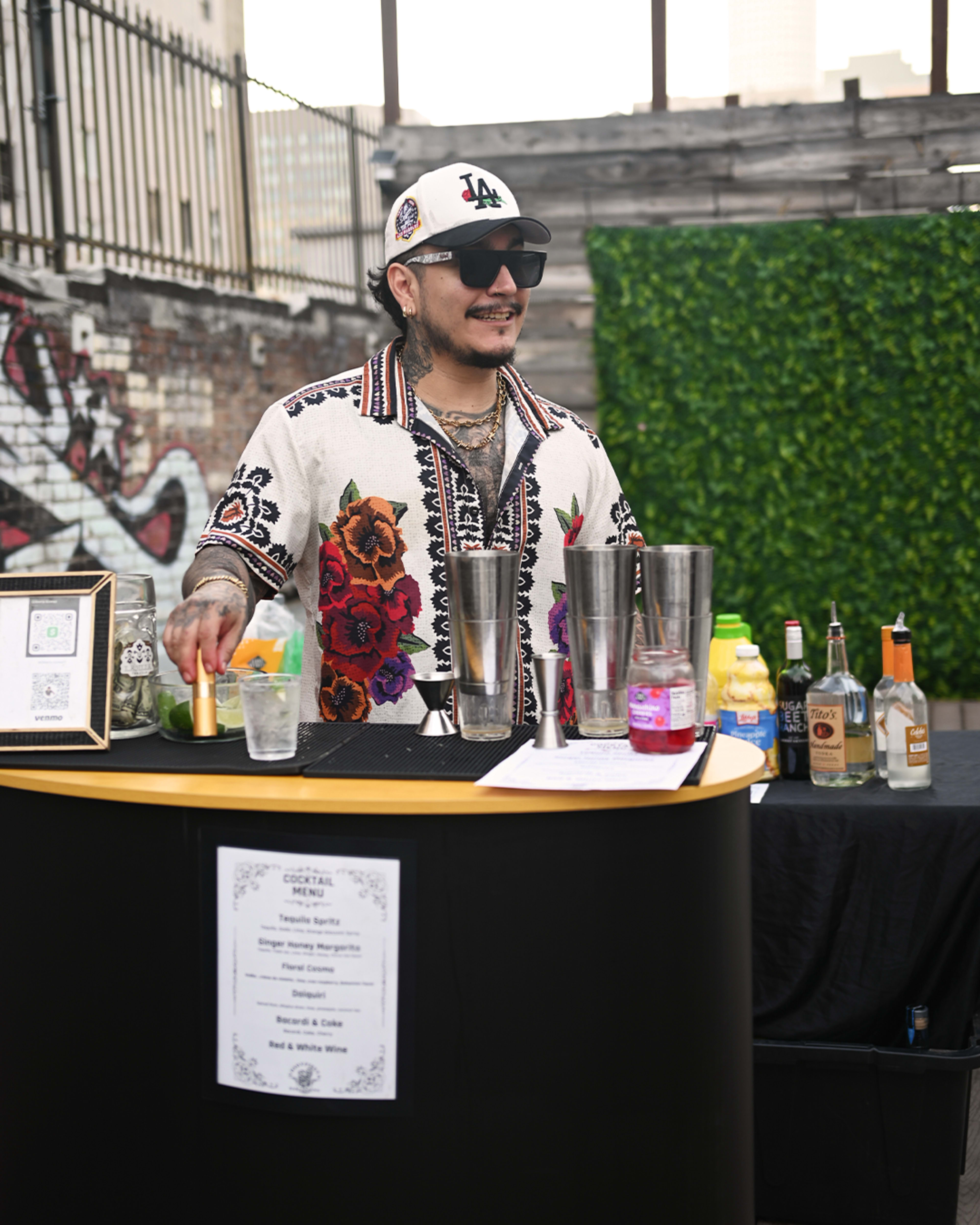 A bartender with tattoos wears a colorful floral shirt and a baseball cap while mixing drinks at a bar.