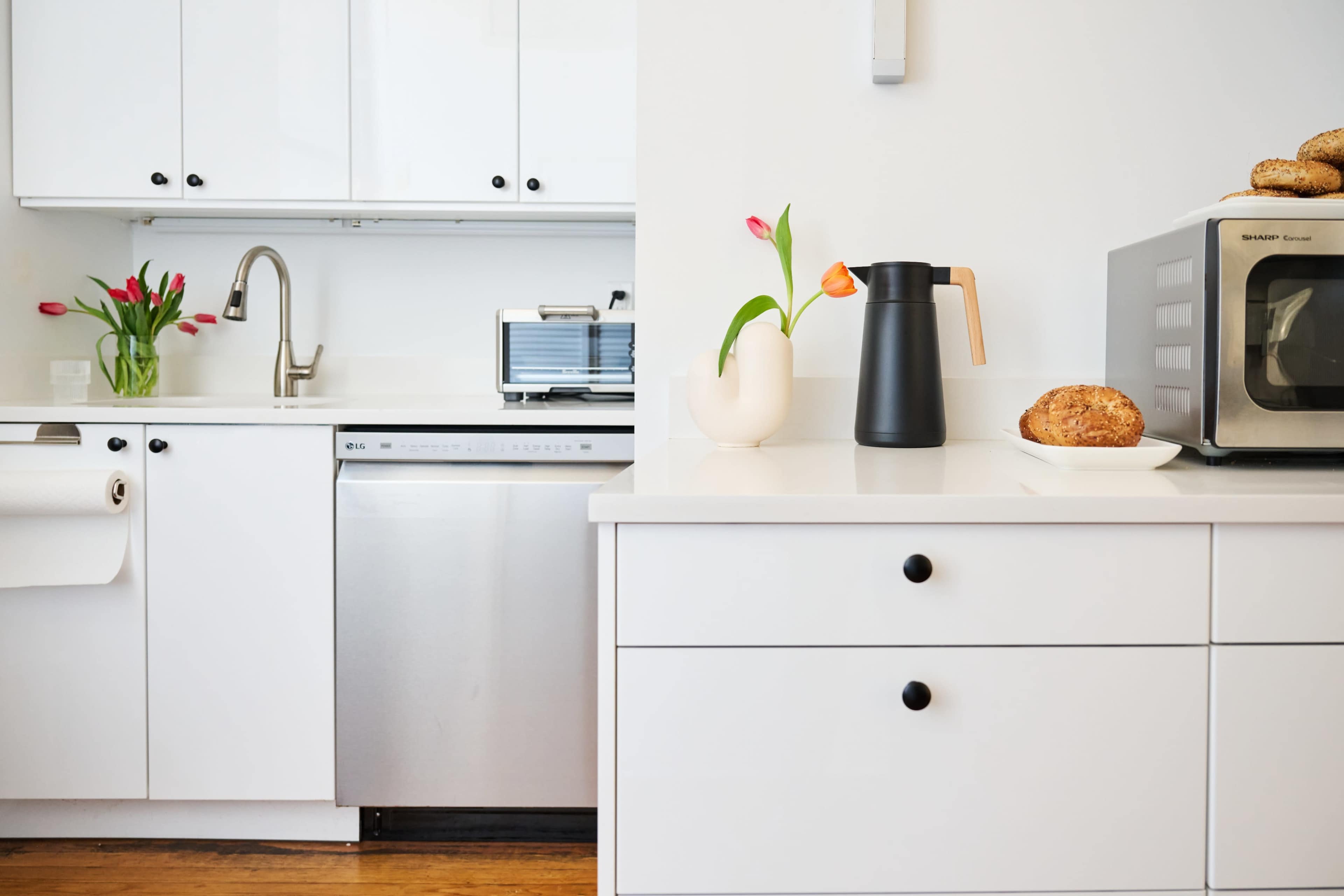 A modern kitchen features white cabinetry, a stainless steel dishwasher, a microwave, and a black kettle on a countertop beside a vase of flowers and a plate of bread.